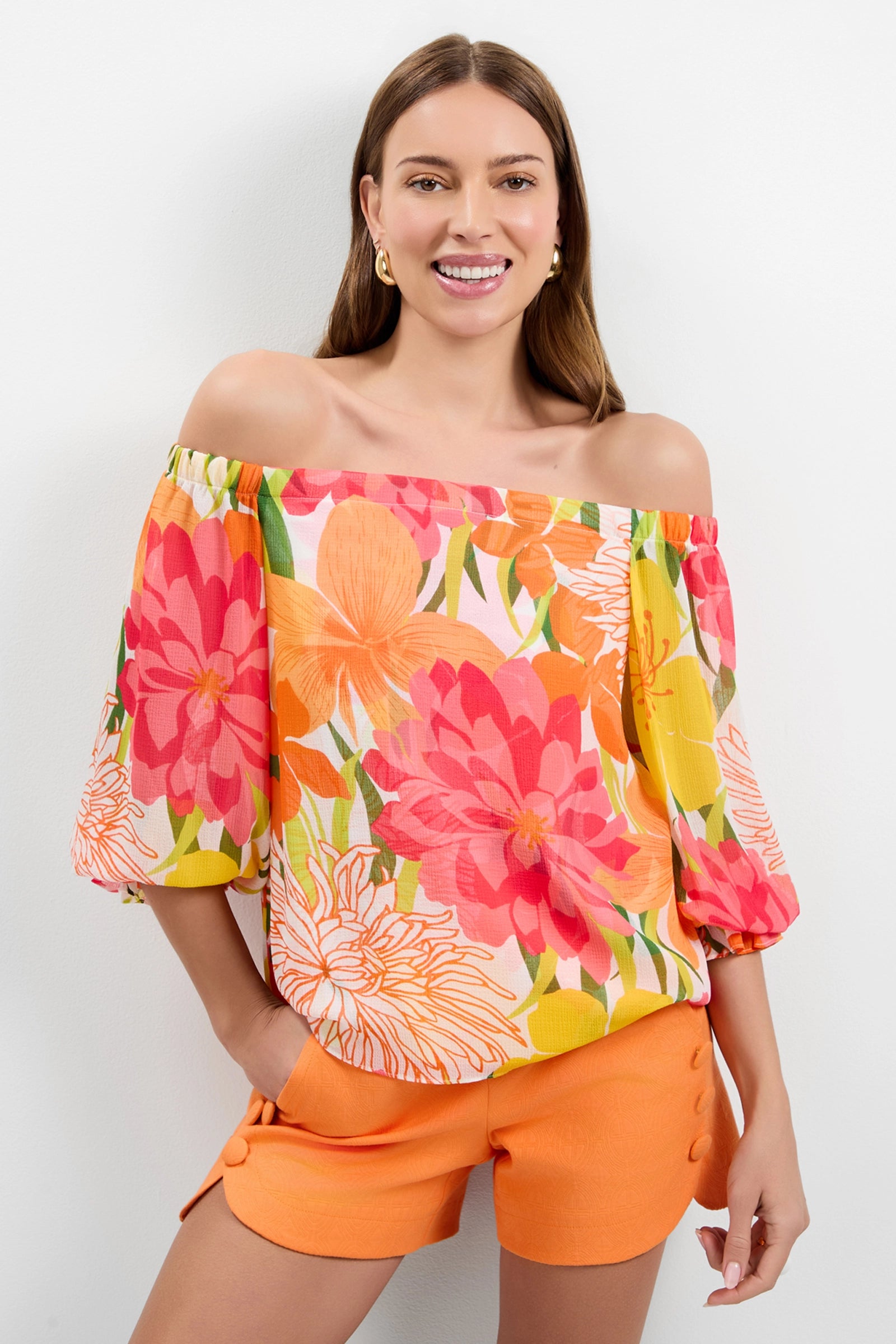 A woman smiles in the Vittoria Off-Shoulder Top, featuring a vibrant pink, orange, and yellow floral pattern, paired with high-waisted orange shorts against a plain white background.