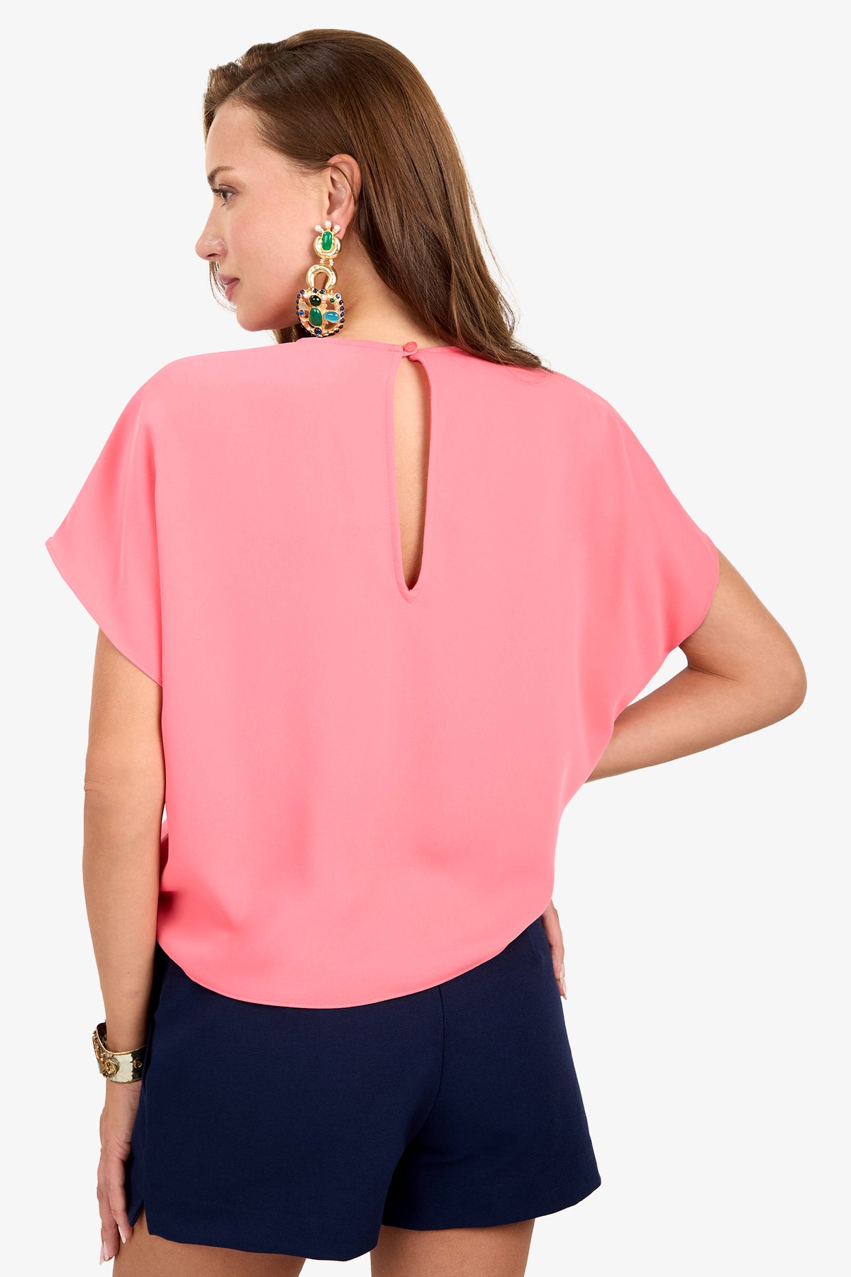 Woman facing away, wearing the Scodelle Top—a loose pink top with a small back keyhole—paired with navy shorts, statement earrings, and a watch, standing against a white background.