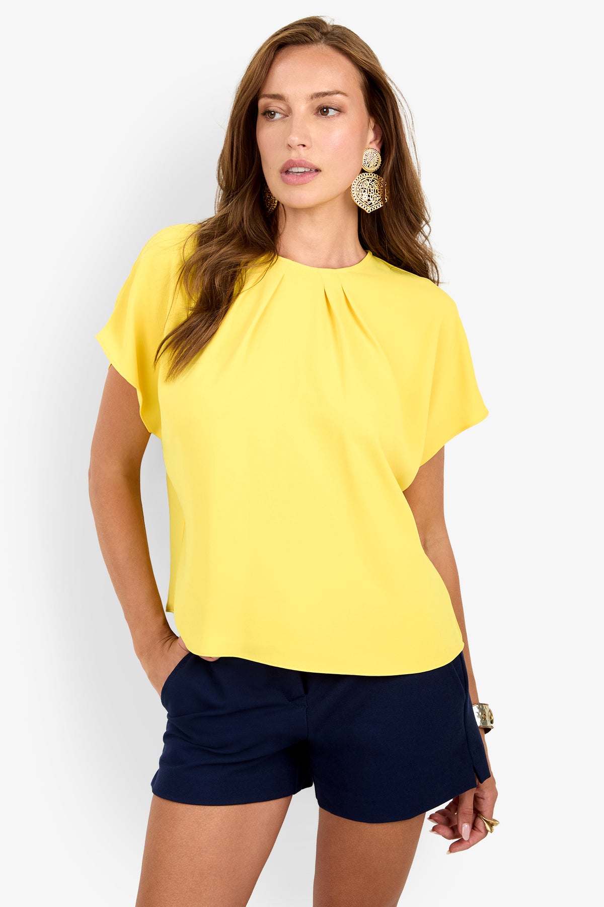 Woman wearing the Scodelle Top, a bright yellow short-sleeve blouse, paired with navy shorts. She stands against a plain white background with one hand in her pocket and a neutral expression.