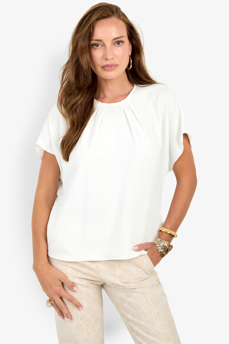 Woman with long brown hair wears the Scodelle Top, a loose white blouse, paired with patterned beige pants, gold bracelets, and hoop earrings against a plain white background.