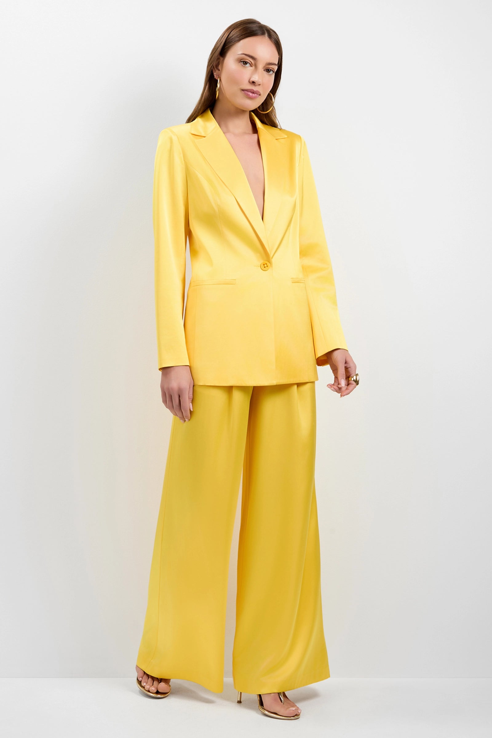 A woman with straight brown hair wears the Giardino Satin Blazer in bright yellow, paired with wide-leg trousers, heels, and gold hoop earrings, standing against a plain background.