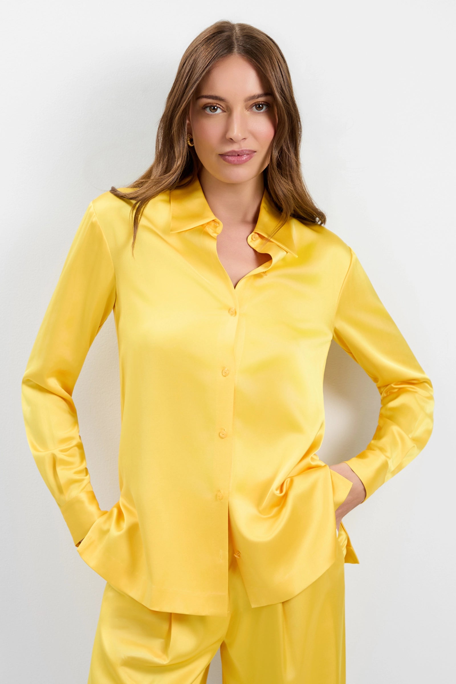A woman with long brown hair wears the Bristlecone Satin Shirt in yellow, paired with matching pants. She stands against a plain white background, hands in her pockets and a slight smile on her face.