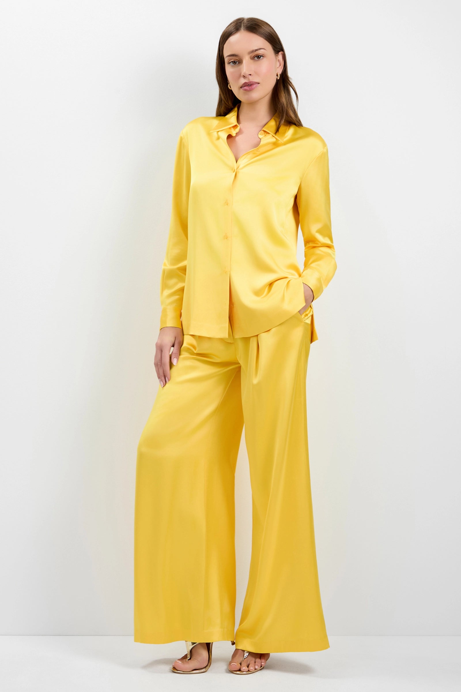 A woman with long brown hair stands barefoot against a plain light background, wearing loose-fitting Pontaccio Satin Pants in bright yellow with wide legs and a matching long-sleeve button-up shirt.