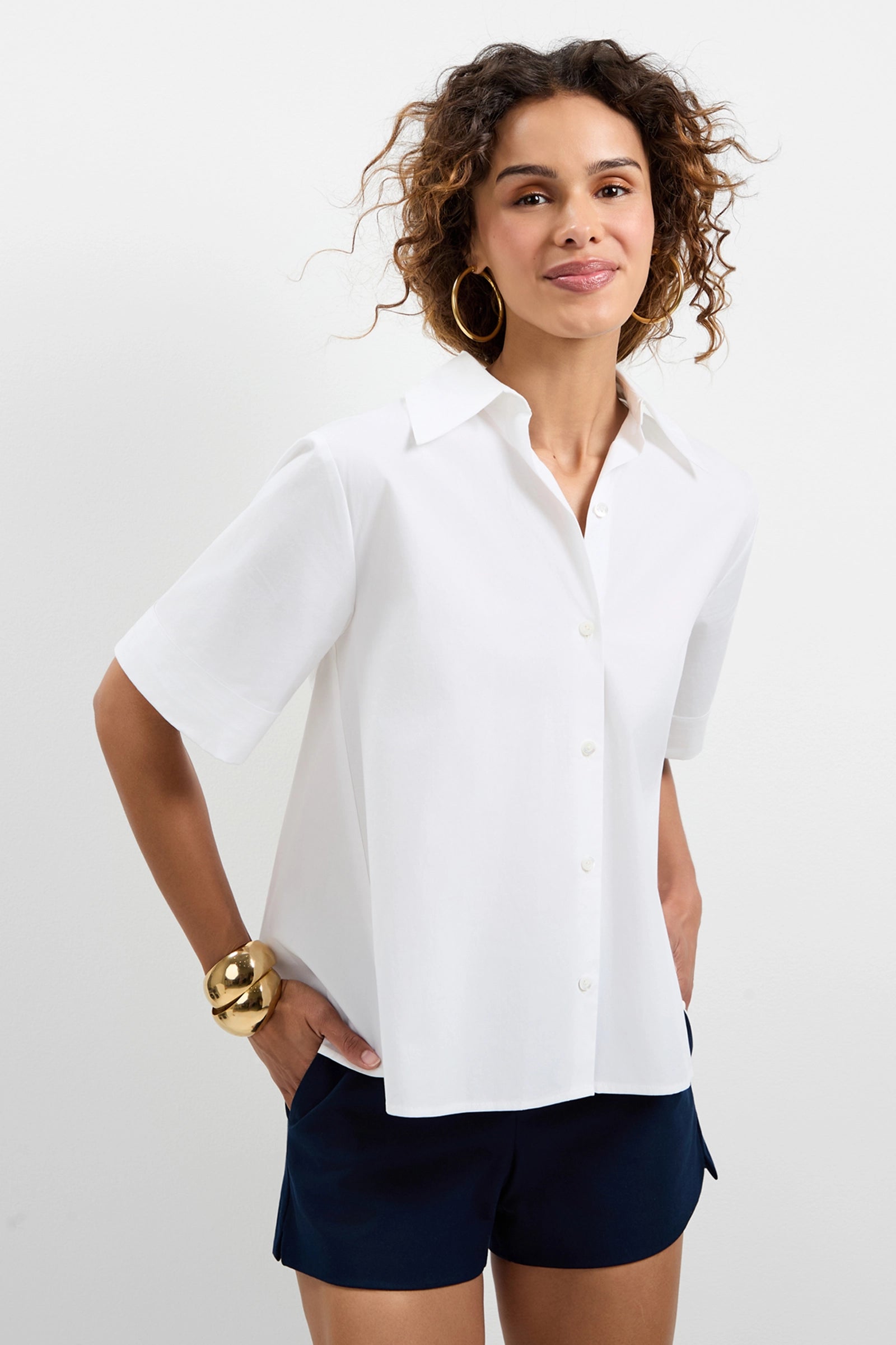 A woman with curly hair wears the Triennale Lace-Up Back Top and dark shorts, smiling against a plain white background. She accessorizes with large hoop earrings and a chunky gold bracelet.