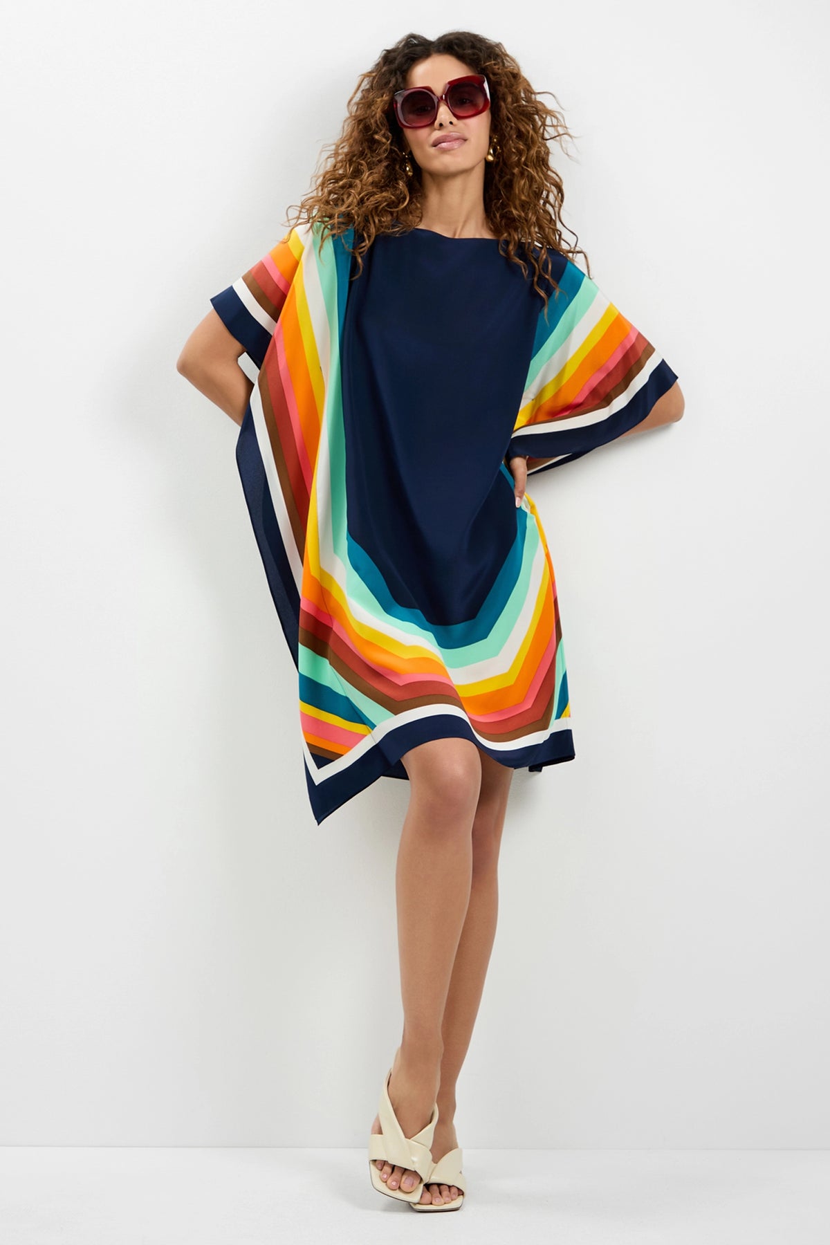 A woman with curly hair wears large pink sunglasses and the Theodora Caftan Dress - Siena, featuring bold diagonal rainbow stripes on navy blue. She poses confidently against a white background, one hand on her hip and legs crossed.