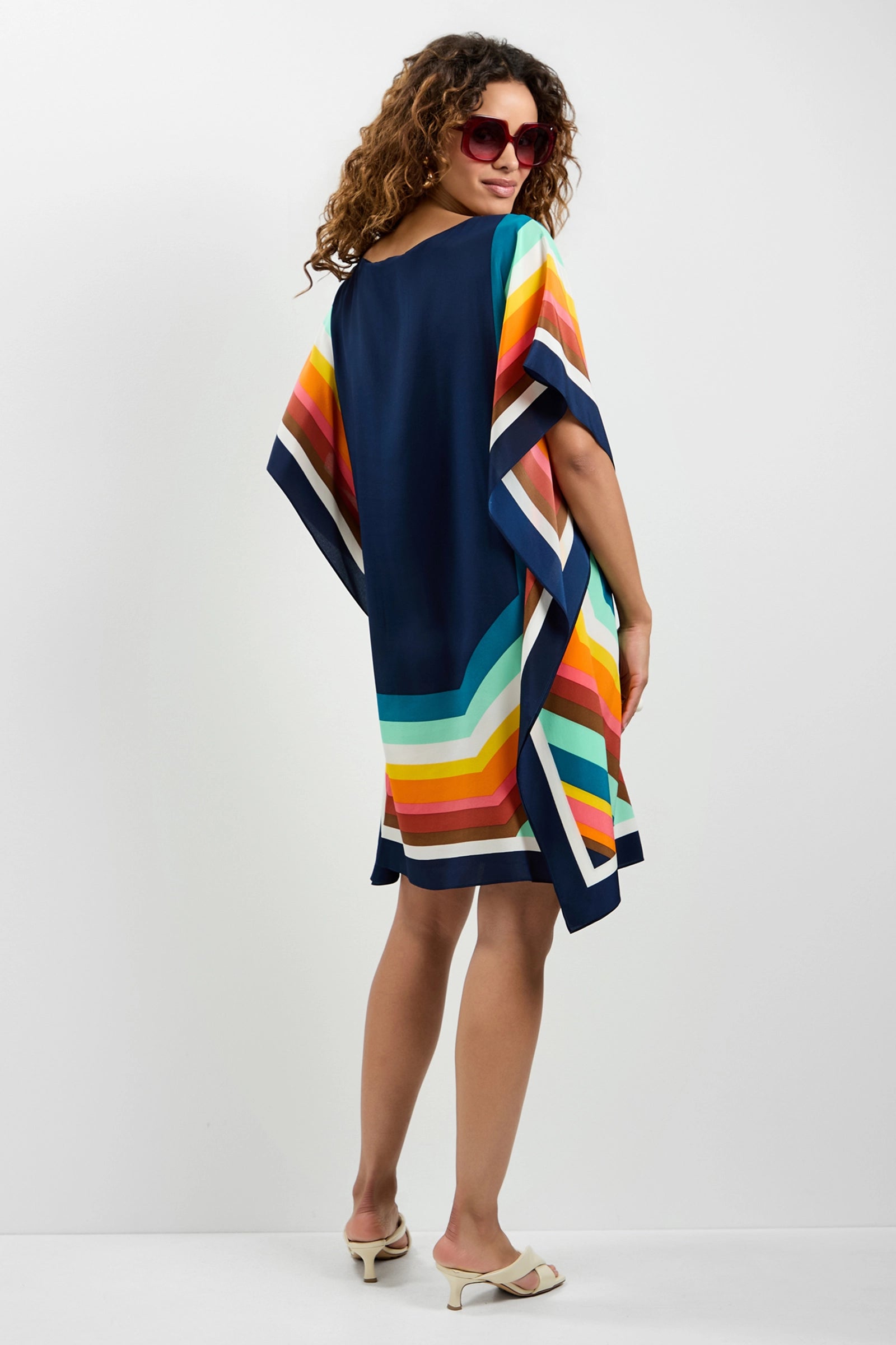 A woman wears the Theodora Caftan Dress - Siena, a loose, knee-length style with colorful horizontal stripes. She looks over her shoulder in red sunglasses and cream heeled sandals against a plain white background.