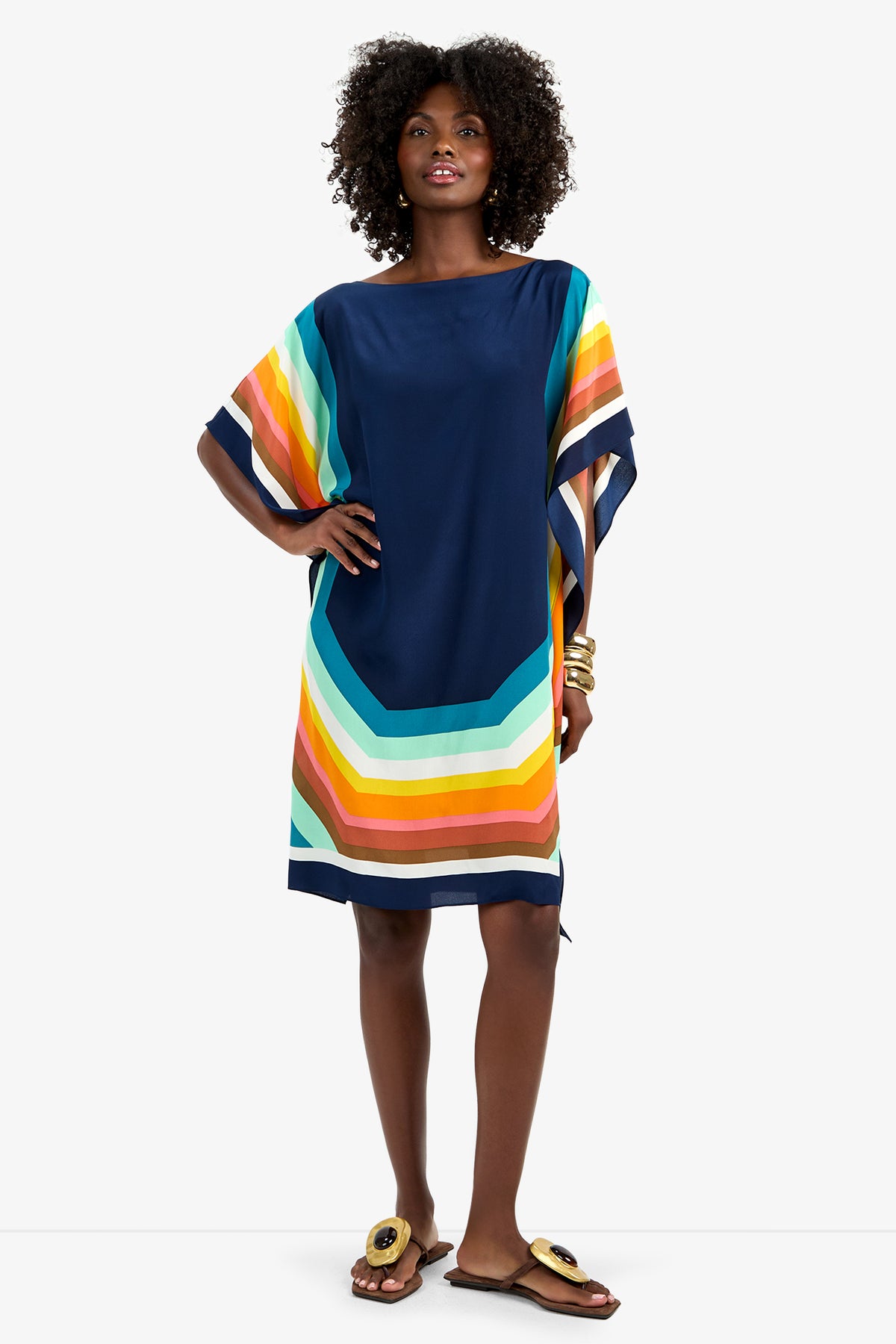 A woman wears Theodora Caftan Dress - Siena, a loose, knee-length navy dress featuring bold orange, yellow, teal, and white stripes on the sleeves and hem. She pairs it with curly hair, gold bracelets, and buckle-detailed sandals.