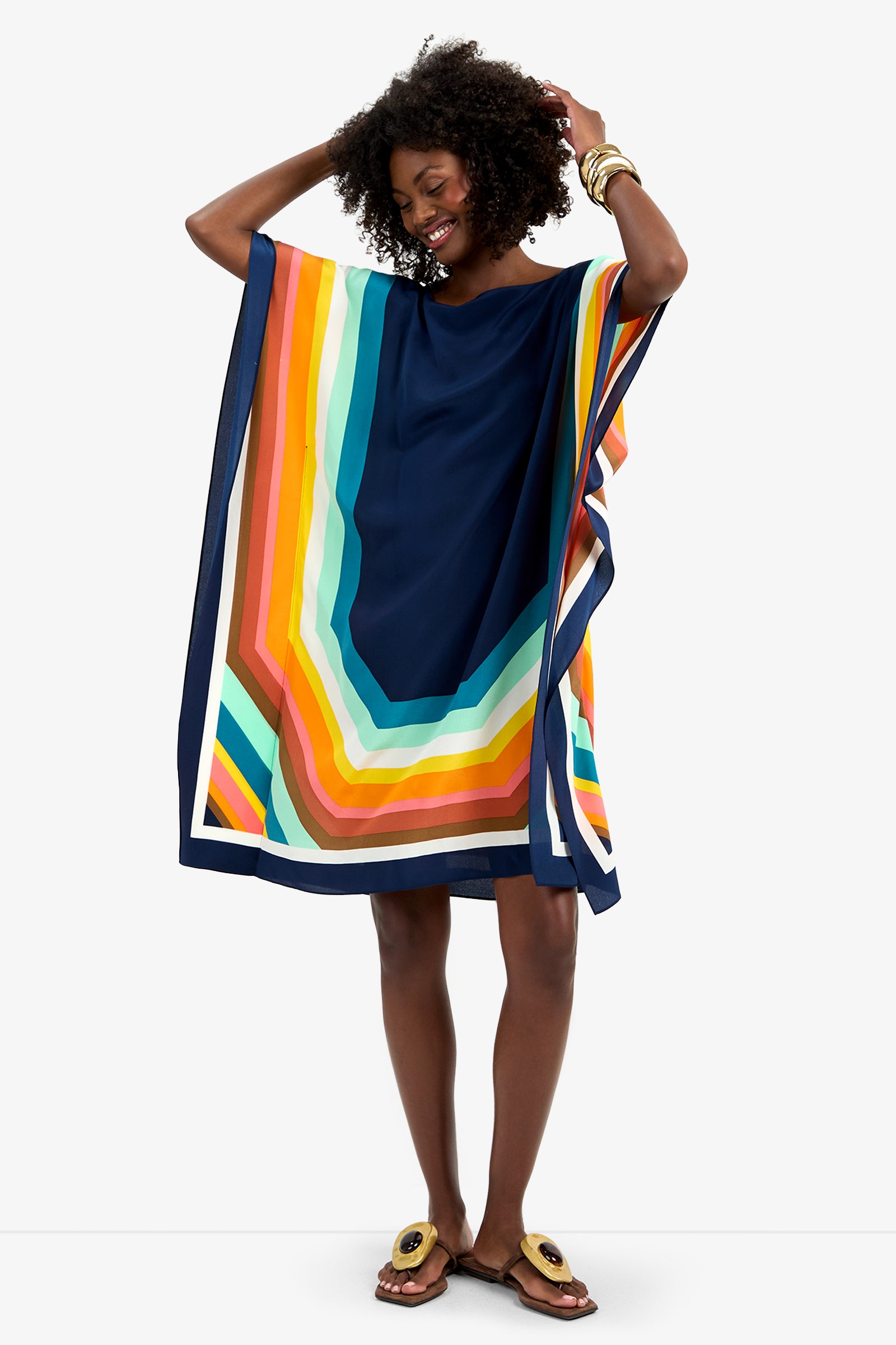 A smiling woman wears the Theodora Caftan Dress - Siena, a loose, colorful geometric-patterned dress with wide sleeves. Barefoot, she crosses her legs, holds her curly hair with both hands, and accessorizes with bangles.