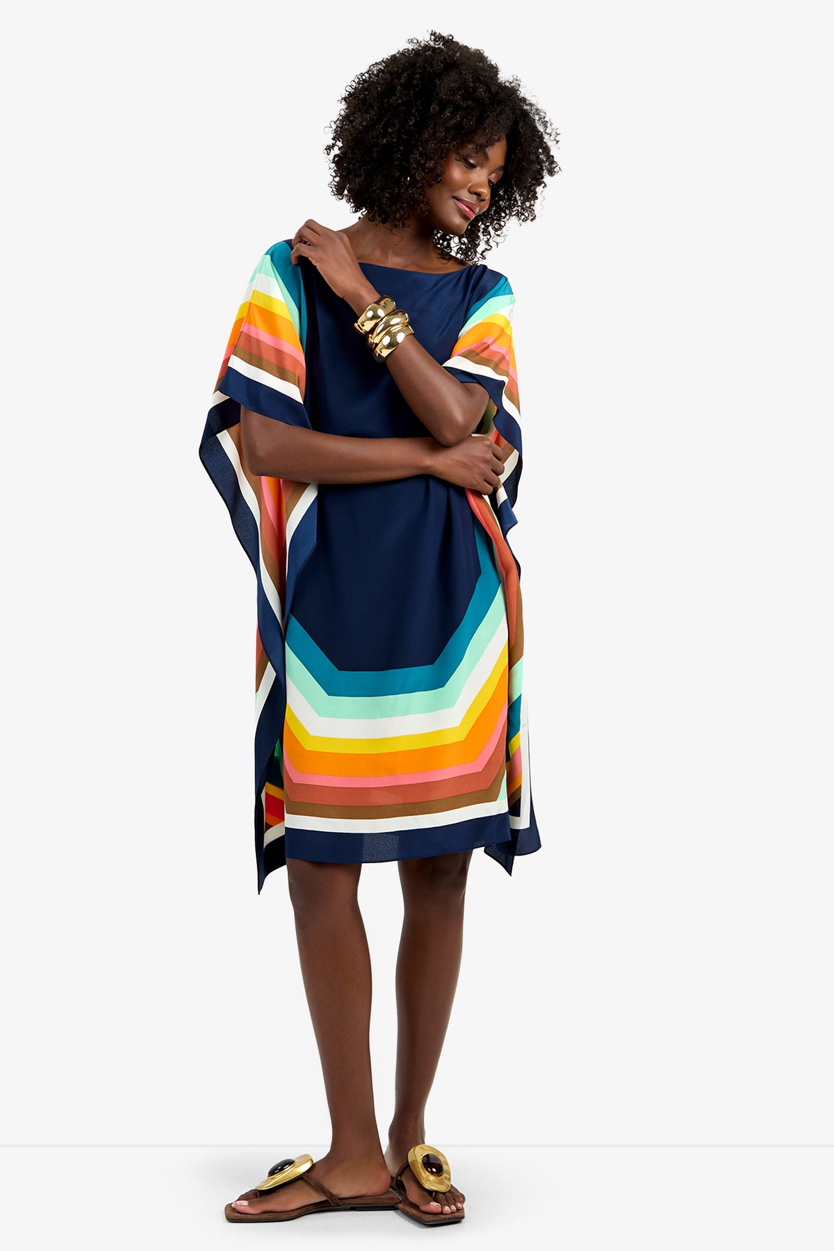 A woman stands smiling against a white background, wearing the Theodora Caftan Dress - Siena, a loose navy blue dress with bold, colorful striped edges, paired with gold bracelets and sandals.