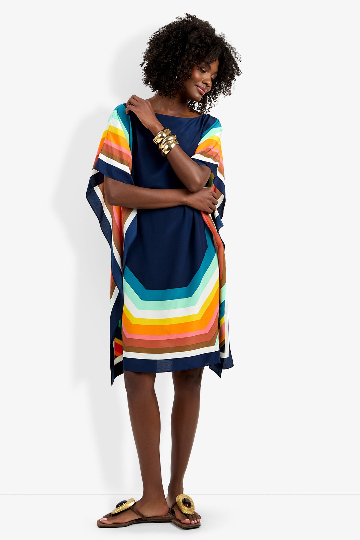 A woman in the Theodora Caftan Dress, a loose navy blue dress with bold colorful stripes, smiles against a plain white background. She accessorizes with gold bangles and sandals, and has curly hair.