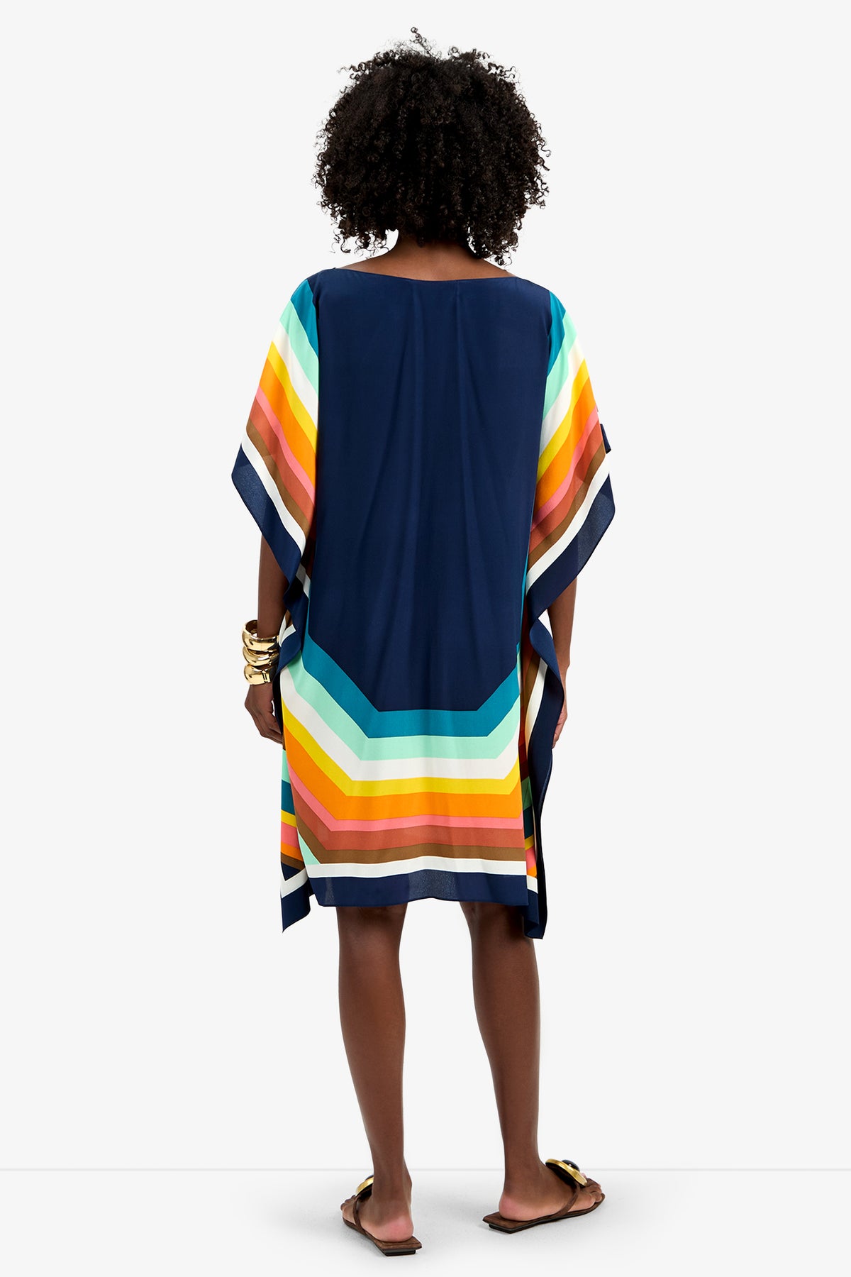 A woman with curly hair faces away, wearing Theodora Caftan Dress - Siena, a loose navy dress featuring wide, colorful horizontal stripes in yellow, orange, green, and blue at the hem and sleeves. She pairs it with sandals and gold bangles.