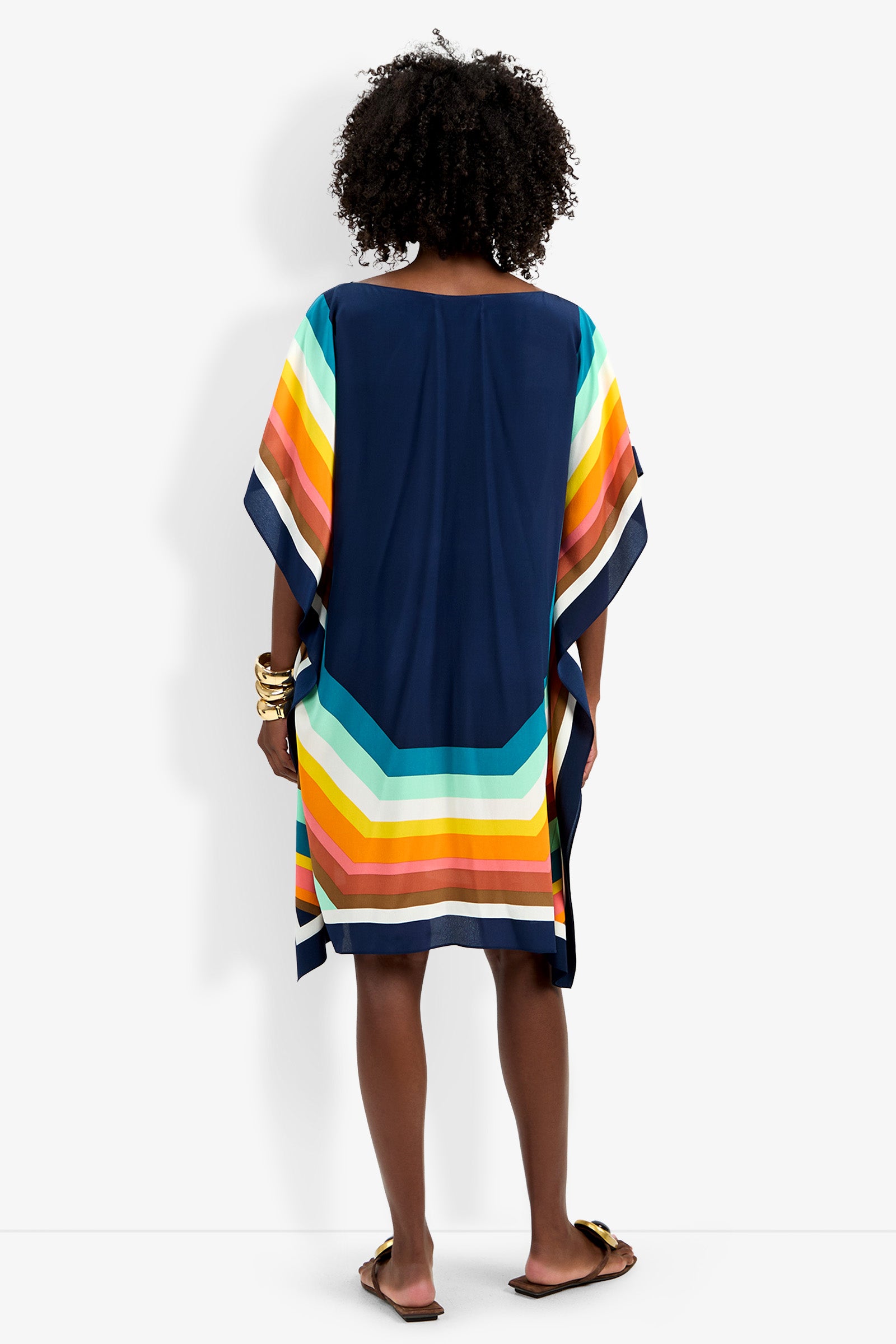 A person with curly hair stands facing away, wearing Theodora Caftan Dress—a loose navy blue dress with colorful yellow, orange, teal, and white stripes near the hem. They pair it with sandals and bangles.