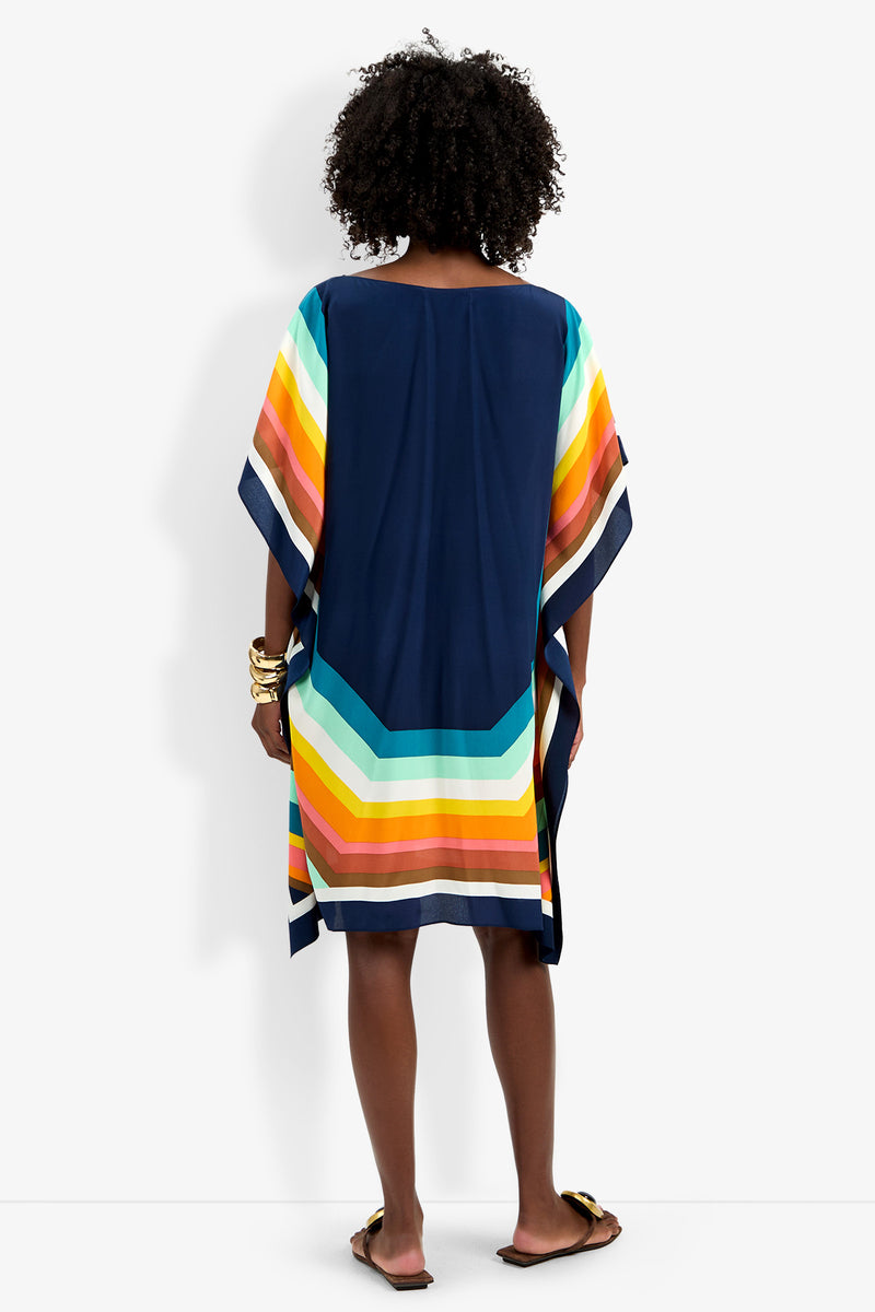 A person with curly hair stands facing away, wearing Theodora Caftan Dress—a loose navy blue dress with colorful yellow, orange, teal, and white stripes near the hem. They pair it with sandals and bangles.