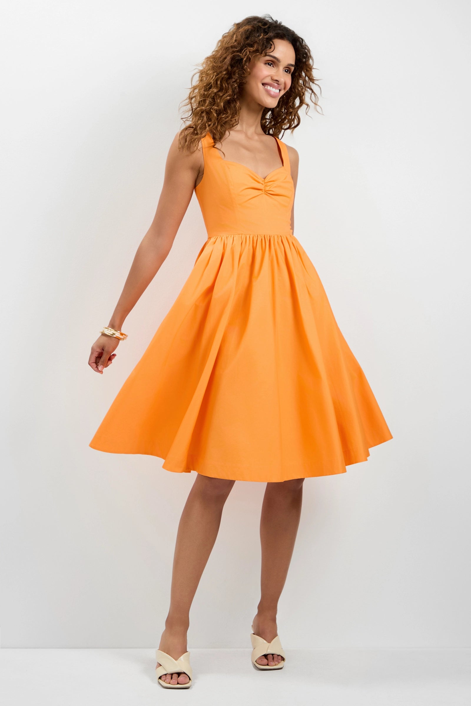 A woman with curly hair smiles against a white background, wearing the Seveso Sweetheart Neck Midi Dress—an orange, sleeveless, fitted-bodice dress with a flared skirt—paired with cream-colored sandals.