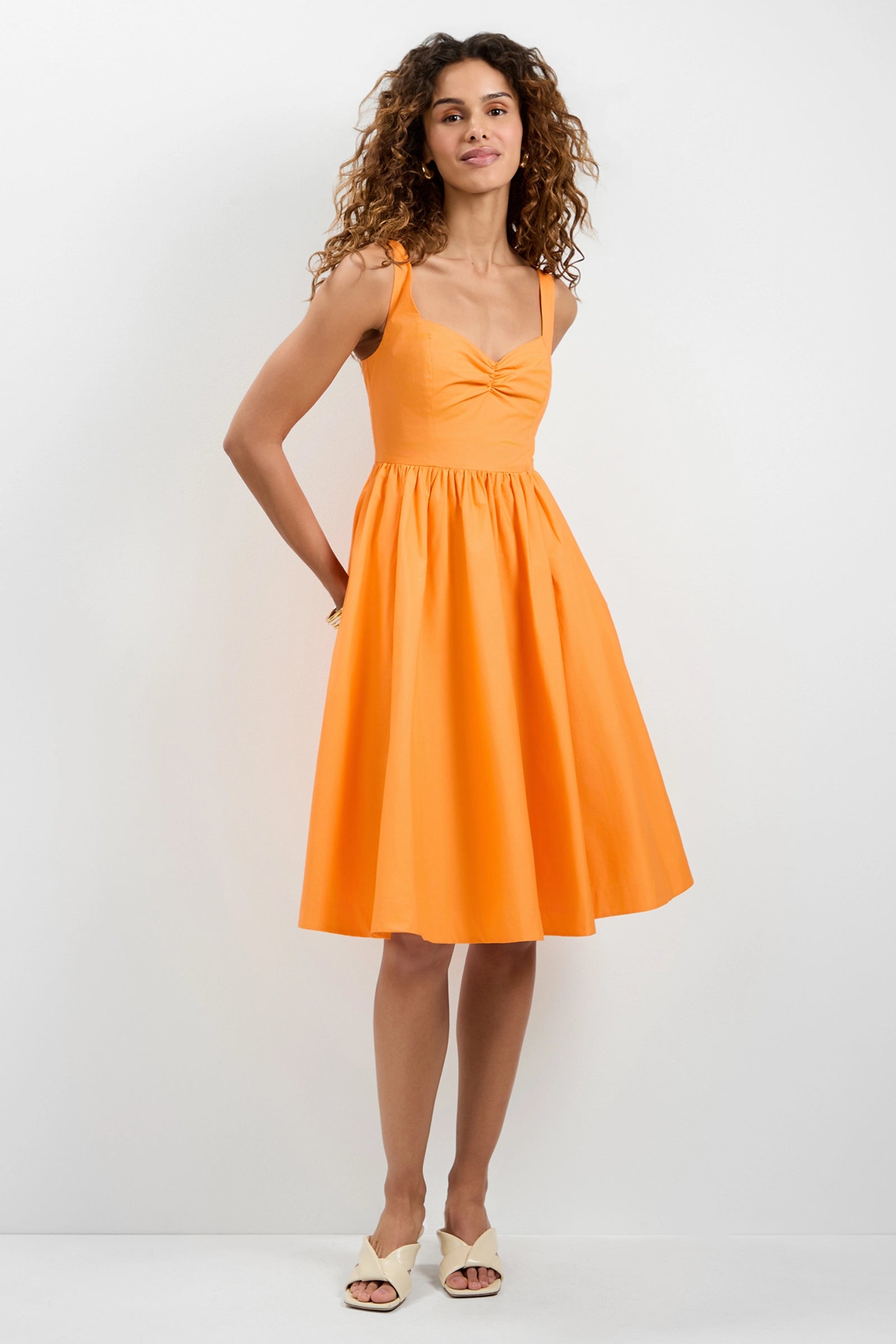 A woman with curly hair stands against a white background wearing the Seveso Sweetheart Neck Midi Dress in bright orange, featuring a sleeveless design, knee-length hem, gathered waist, and front knot detail. She pairs it with white open-toe shoes.