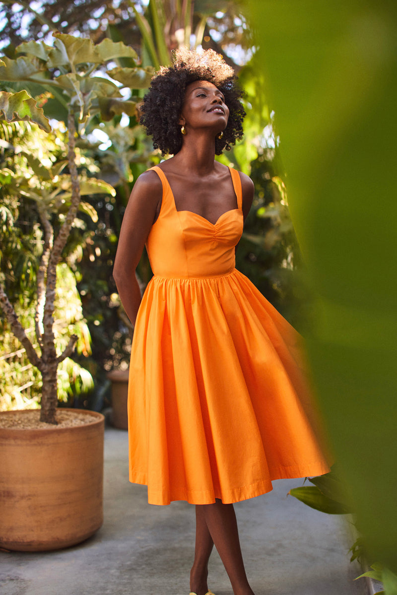 A woman stands among green plants, wearing the Seveso Sweetheart Neck Midi Dress in bright orange. Sunlight highlights her face and curly hair as she looks upward with a relaxed expression.