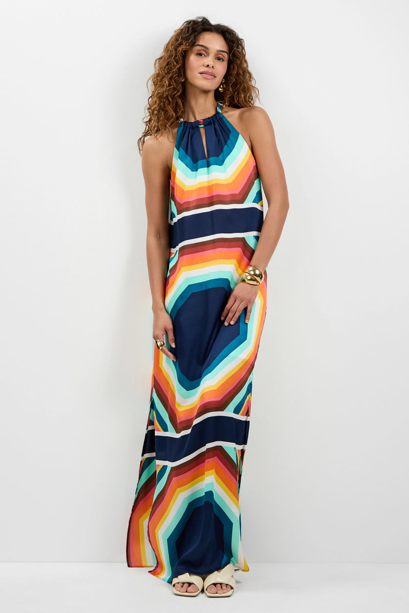 A woman with curly hair wears the Emilo Sleeveless Maxi Dress, featuring bold, colorful geometric stripes, paired with white sandals and gold bangles. She stands against a plain white background.