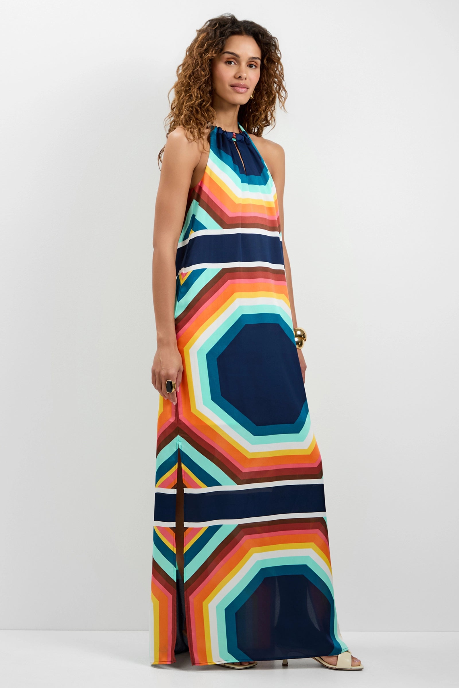 A woman with curly hair models the Emilo Sleeveless Maxi Dress, a floor-length gown with bold, colorful geometric stripes and octagonal patterns, standing against a plain white background.