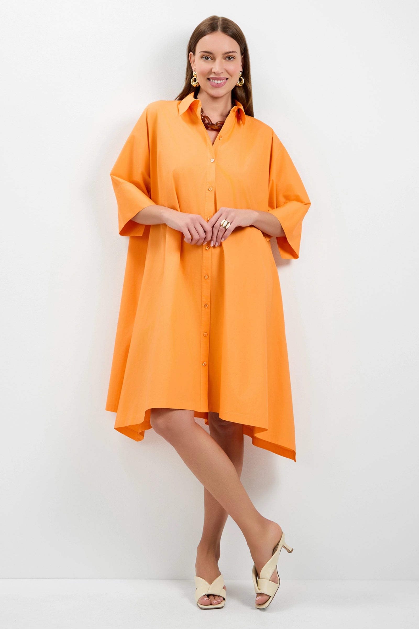 A woman smiles against a white wall, wearing the oversized Mirabello Shirt Dress in orange with wide sleeves, gold jewelry accents, and cream heeled sandals.
