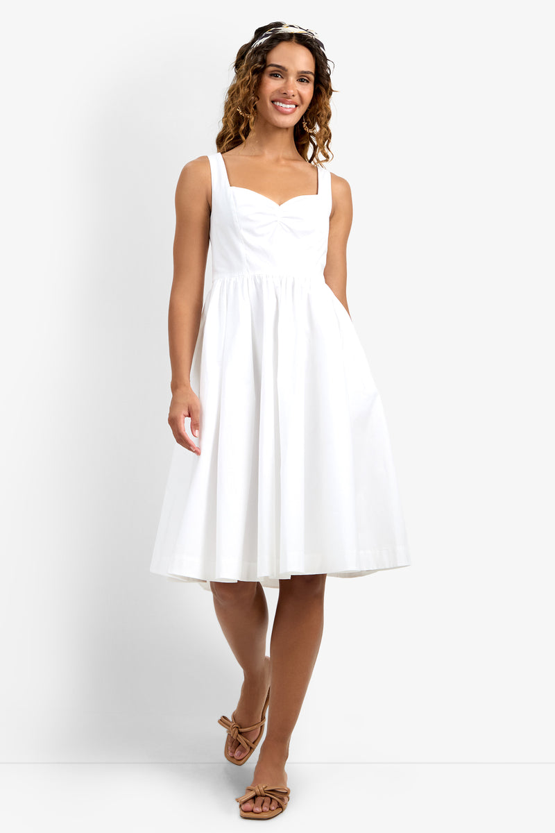 A woman in the Mirabello Sweetheart Neck Midi Dress—sleeveless, white, knee-length with a fitted bodice and flared skirt—smiles in sandals and a headband against a plain light background.