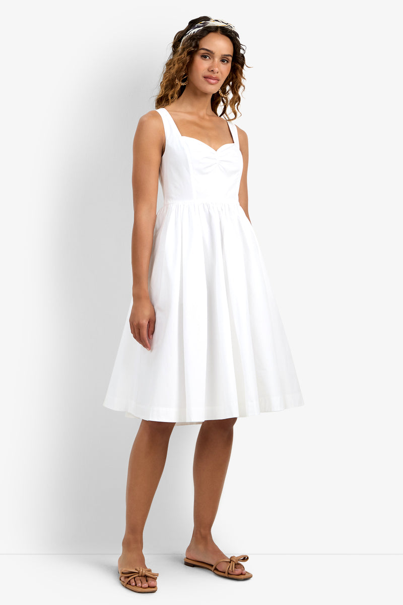 A woman wearing the Mirabello Sweetheart Neck Midi Dress, a sleeveless, knee-length white dress with a fitted bodice and flared skirt, paired with brown sandals and a headband, stands against a plain white background.