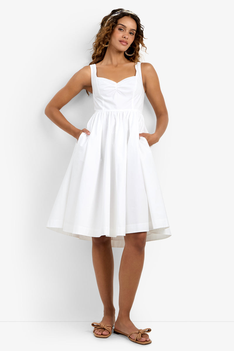 A woman wearing the Mirabello Sweetheart Neck Midi Dress, a sleeveless white dress with a fitted bodice, flared skirt, and pockets, paired with flat brown sandals.