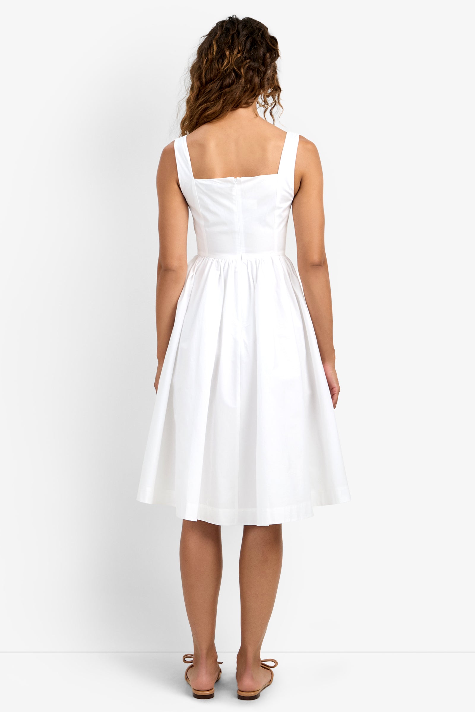 Woman with long curly hair, seen from behind, models the Mirabello Sweetheart Neck Midi Dress in white against a plain background. She wears sandals and the dress is sleeveless and knee-length.