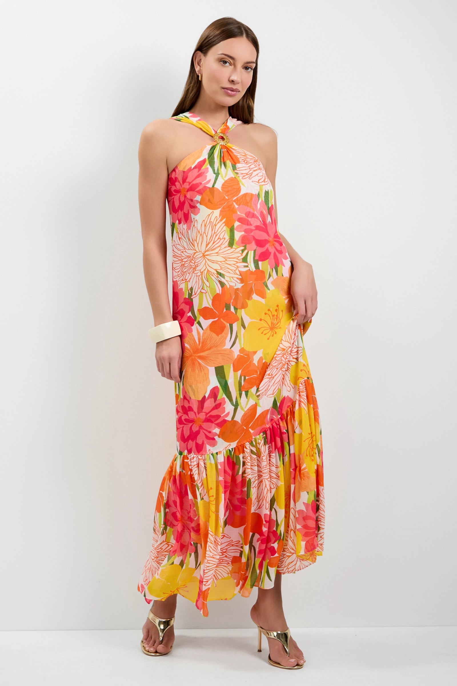 A woman with straight brown hair wears the Navigli Sleeveless Maxi Dress, featuring colorful pink, orange, yellow, and green floral patterns. She pairs it with heeled sandals and a white bangle against a white background.