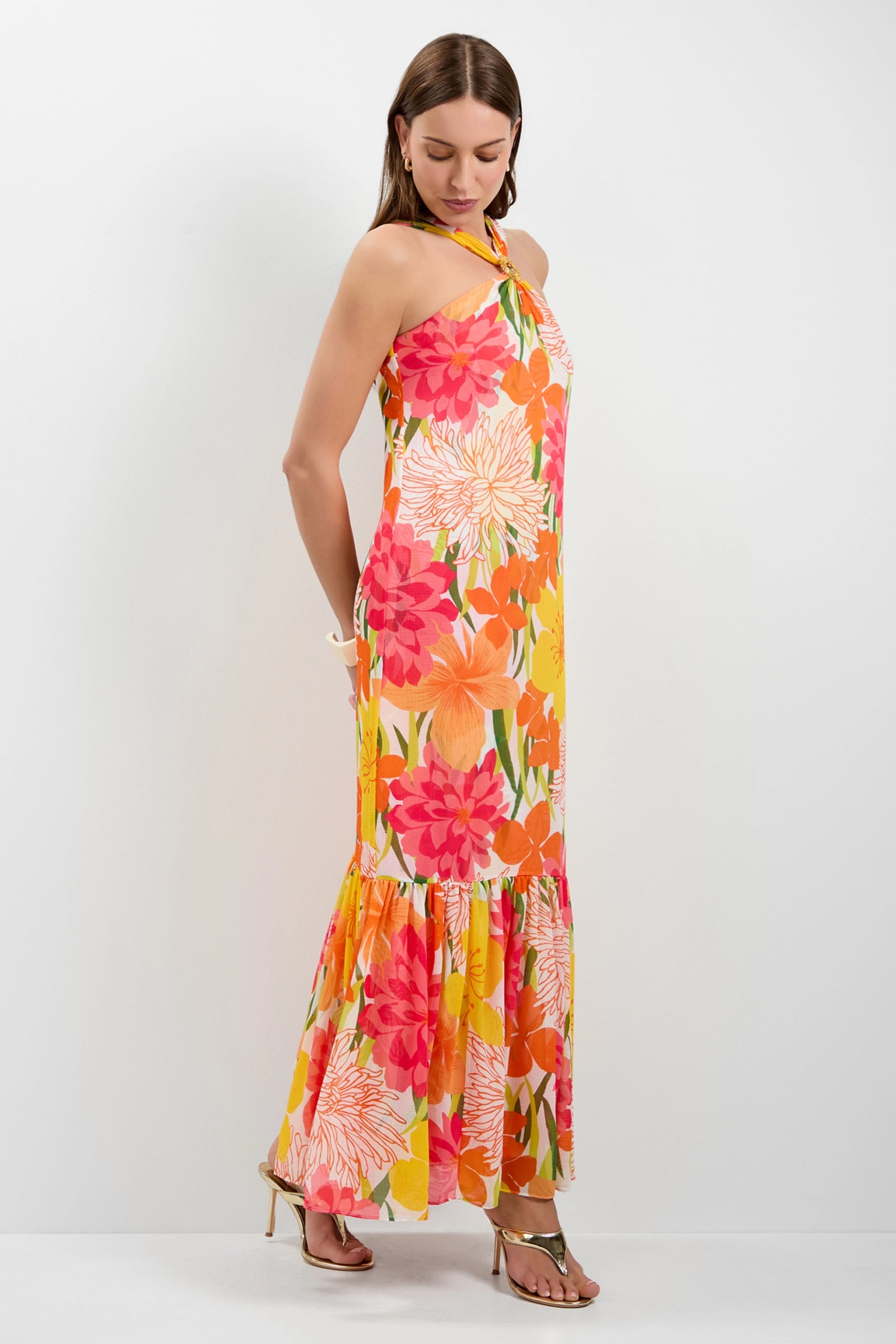 A woman wearing the Navigli Sleeveless Maxi Dress, featuring a bright floral print in pink, orange, and yellow, with strappy heeled sandals stands against a plain white background.