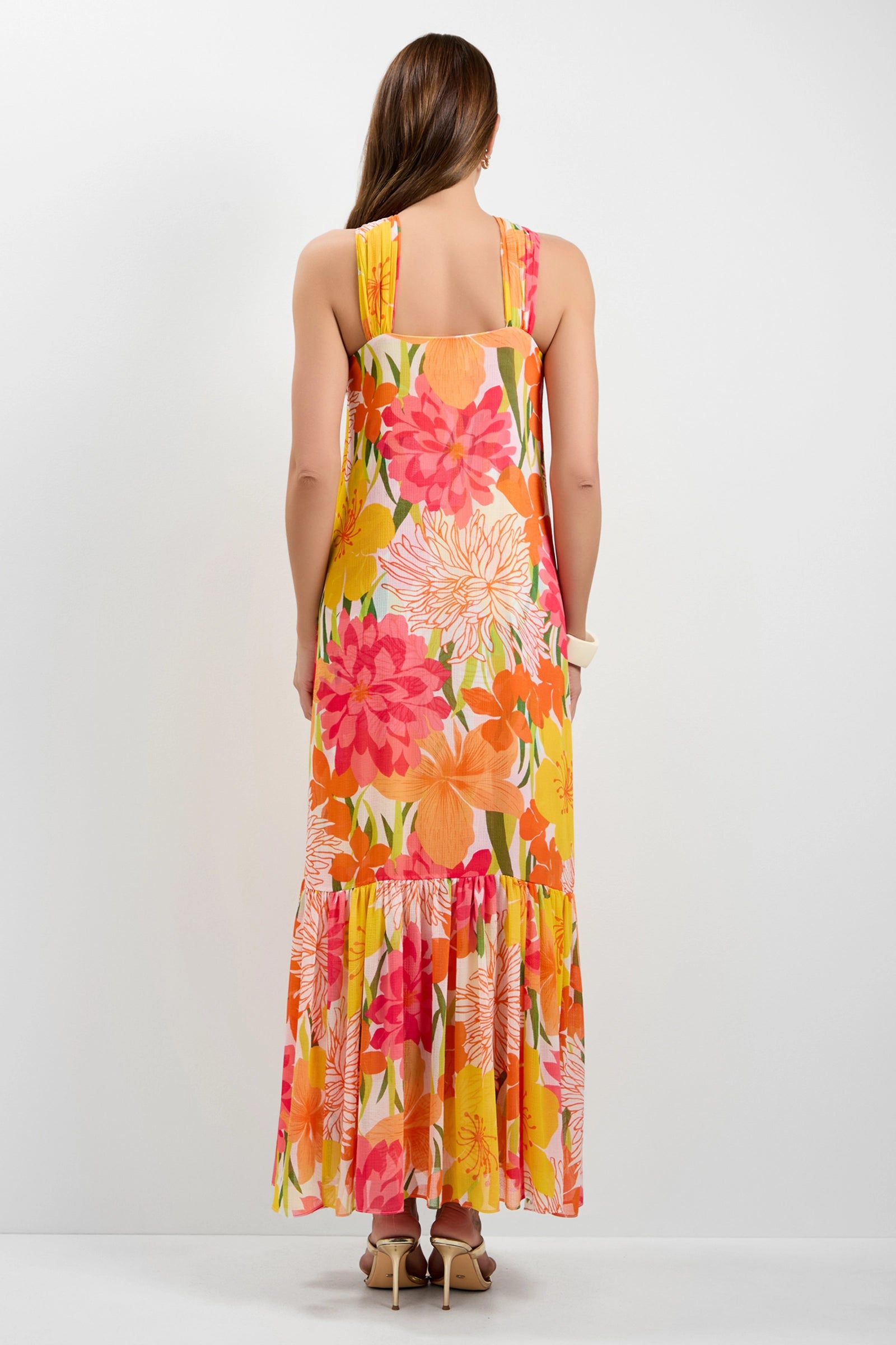 A woman with long brown hair, wearing beige heels, stands facing away in the Navigli Sleeveless Maxi Dress featuring a vibrant floral pattern in pink, orange, and yellow.