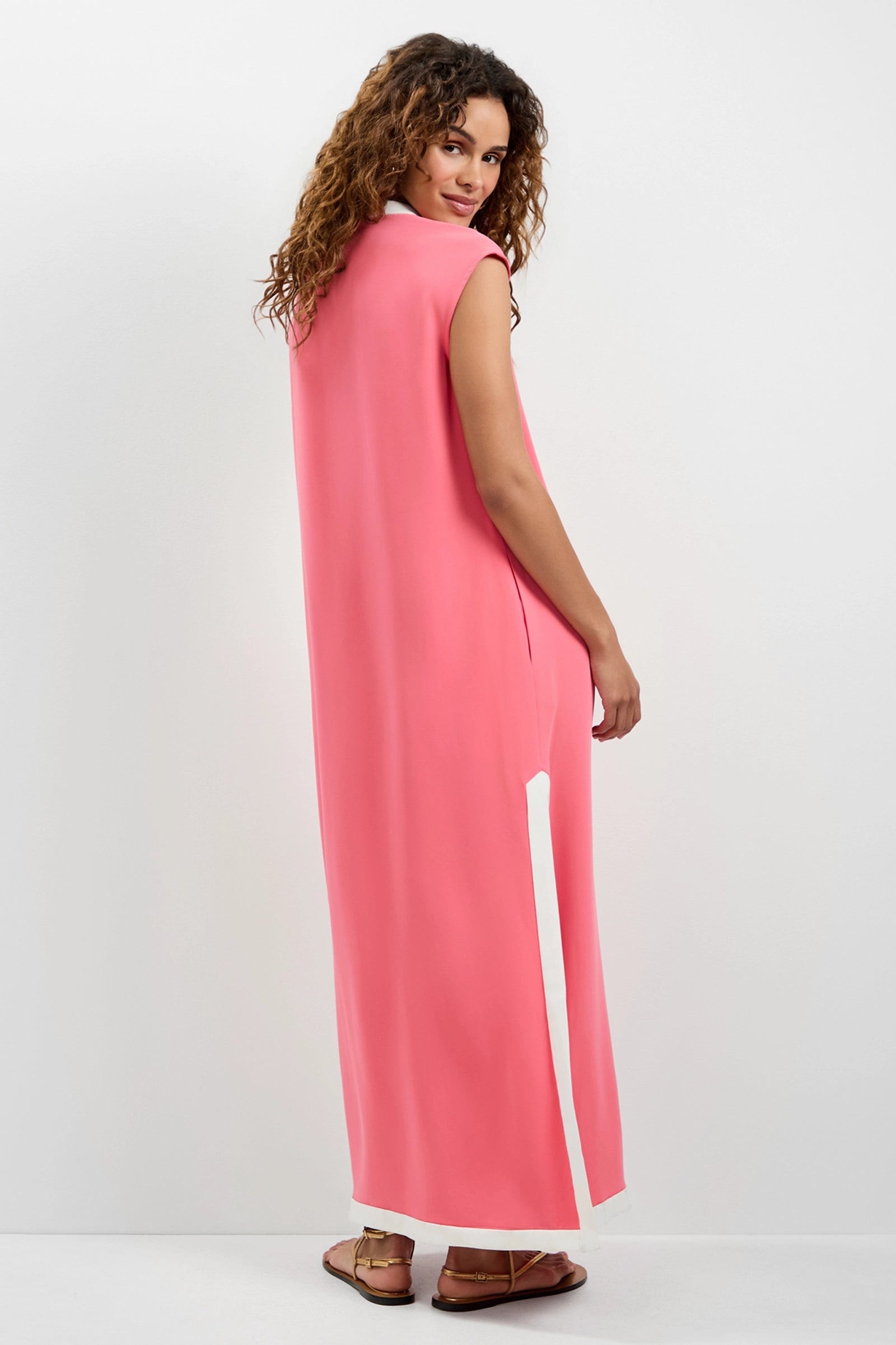 A woman with curly hair, wearing the Nuvola Zip-Front Midi Dress in pink with white accents and brown sandals, stands facing away and looks over her shoulder against a plain white background.