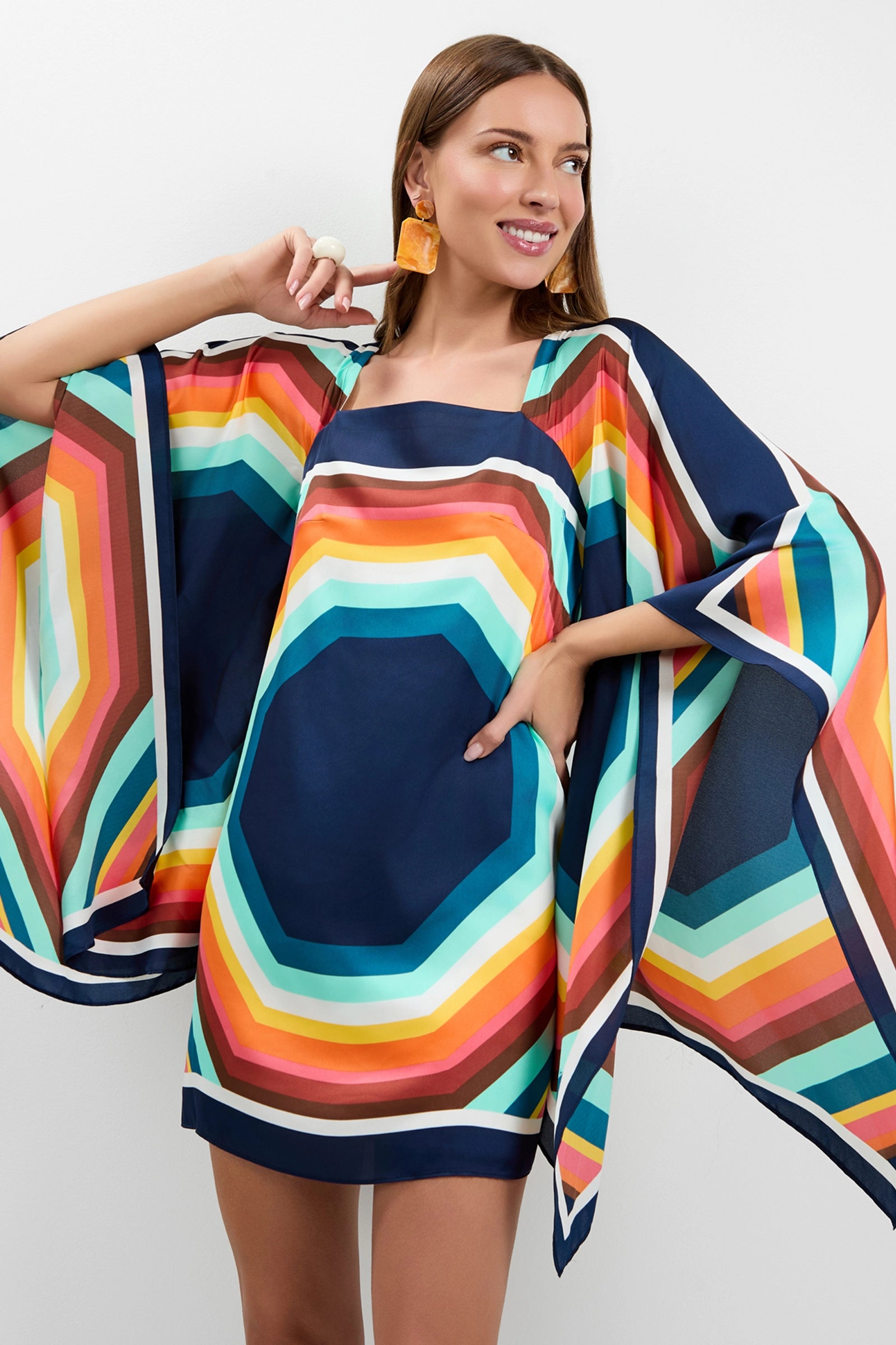A smiling woman poses in the Aqualane Side Flounce Dress, featuring wide sleeves and bold geometric stripes in blue, orange, yellow, and green. She accessorizes with statement earrings and a white ring.