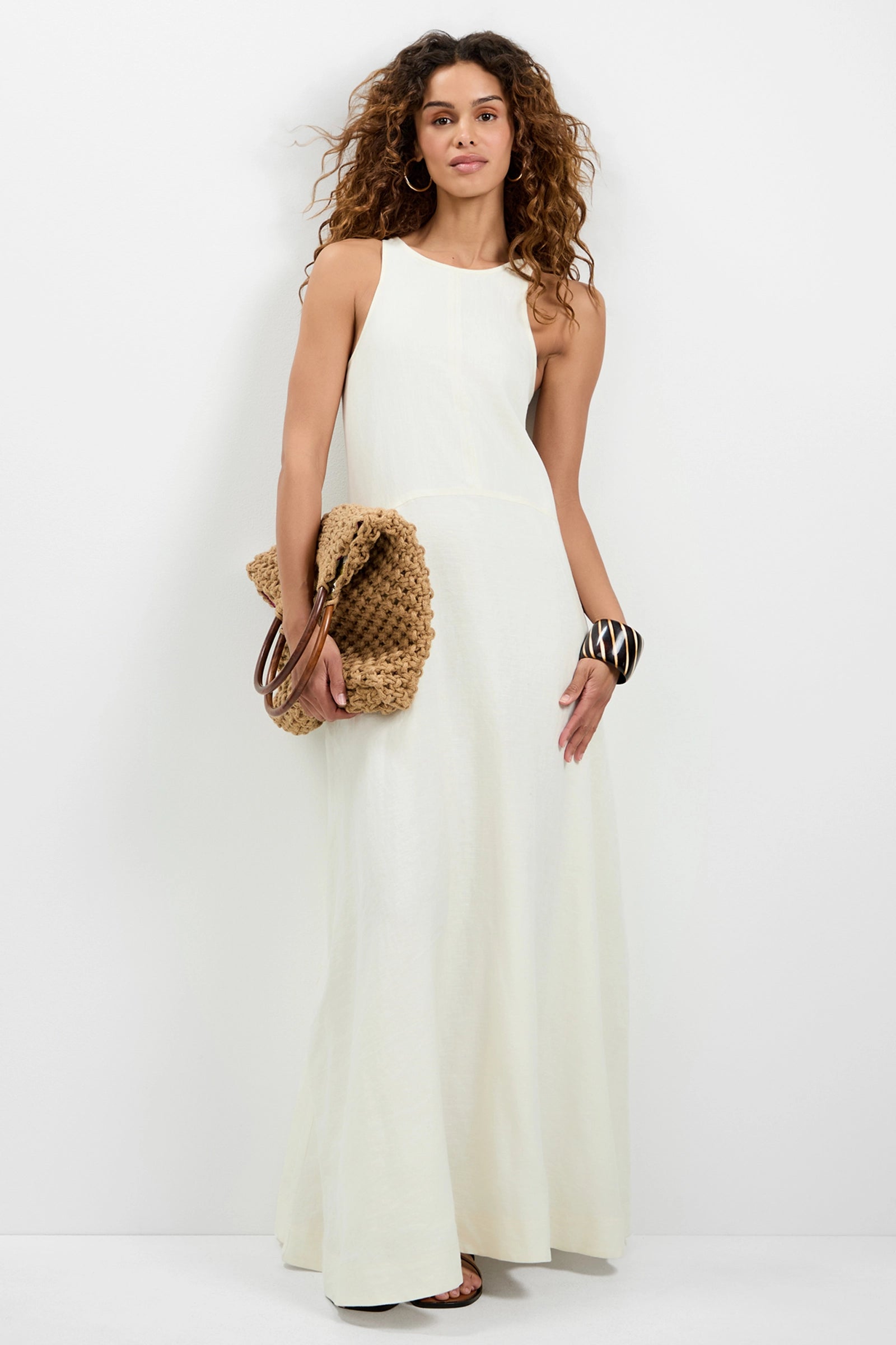 A woman with curly hair wears the white Pichi 2 Maxi Dress, holding a woven tan clutch. She accessorizes with hoop earrings, bangles, and sandals while posing against a plain white background.