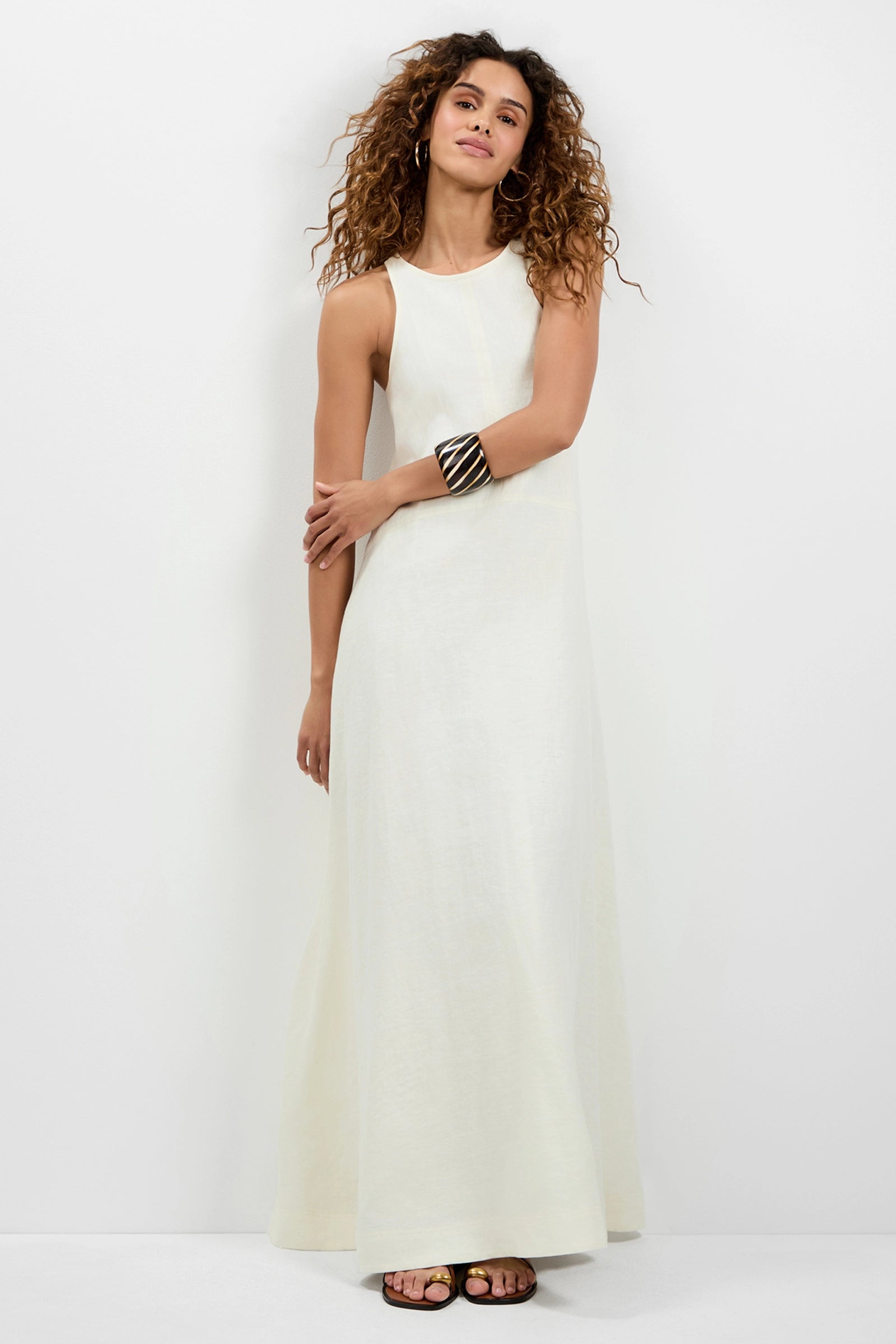 A woman with curly hair wears the Pichi 2 Maxi Dress—sleeveless, long, and white—paired with sandals and minimal jewelry as she stands in a relaxed pose against a plain white background.