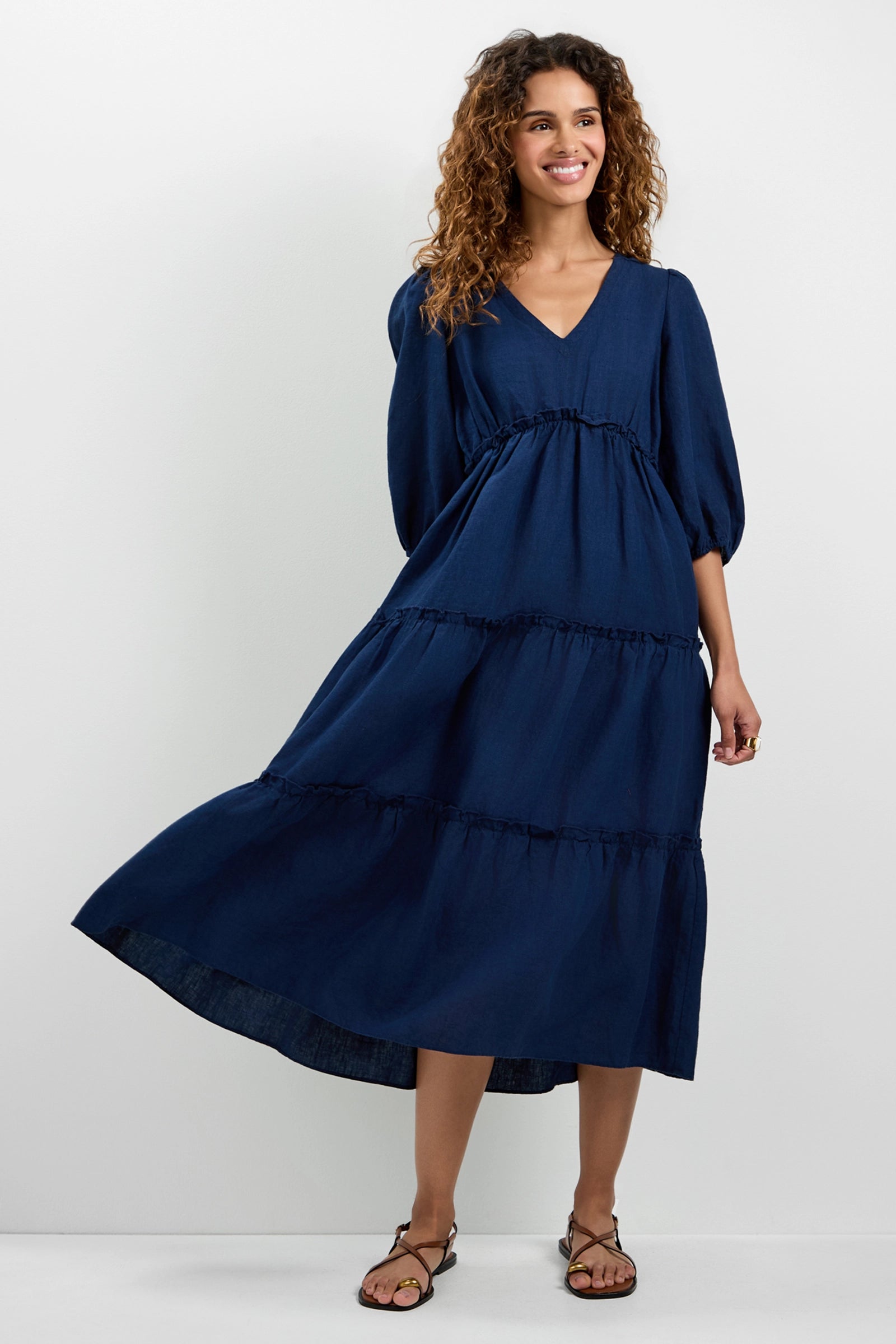 A woman with curly hair wears the Calm Tiered Ruffle Midi Dress in navy blue, featuring elbow-length puff sleeves and a V-neckline. She smiles against a plain light background, holding out one side of her dress and wearing sandals.