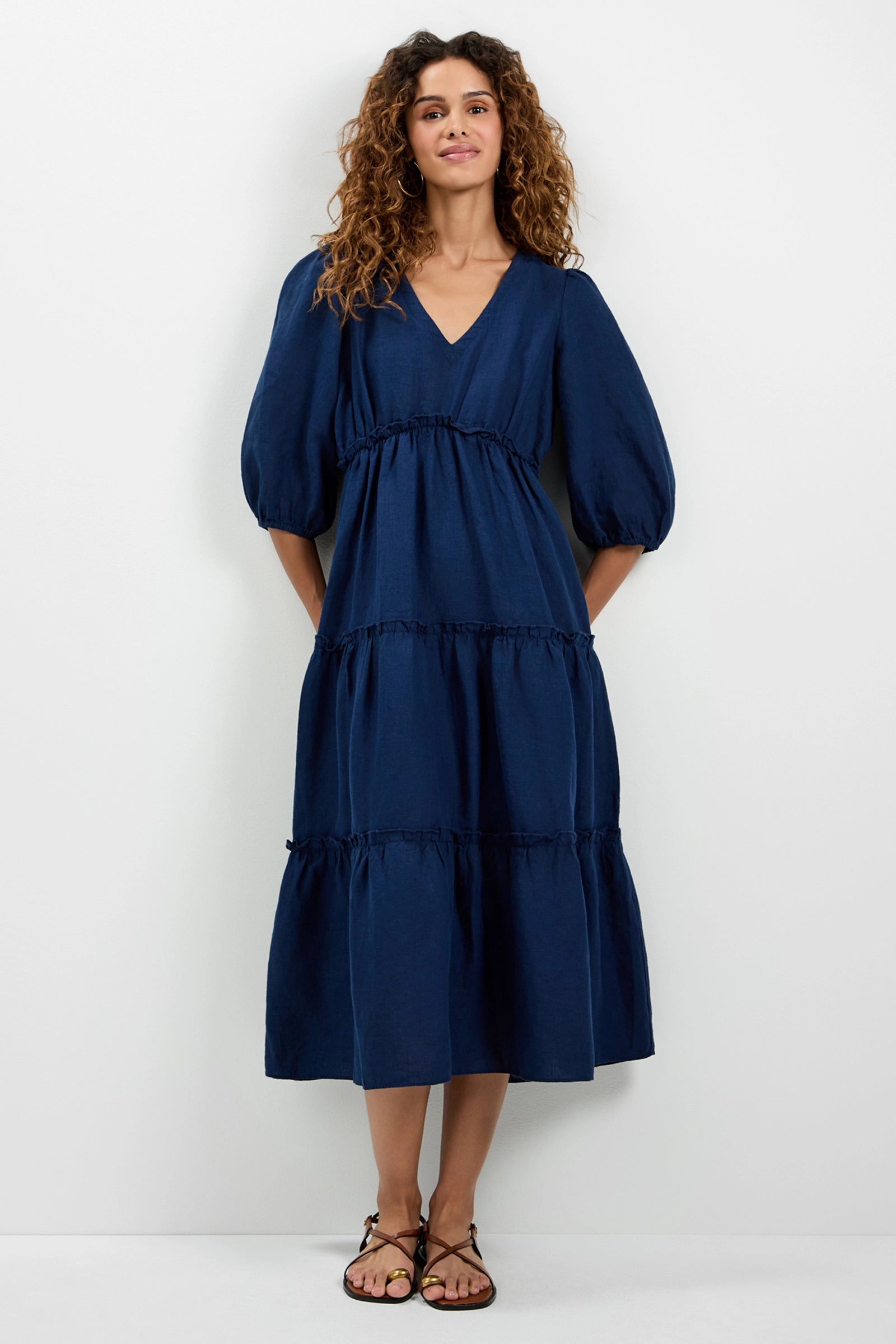 A woman with curly hair smiles as she models the Calm Tiered Ruffle Midi Dress in Ink, featuring puffed sleeves and a V-neckline. She stands against a white background and wears black sandals.