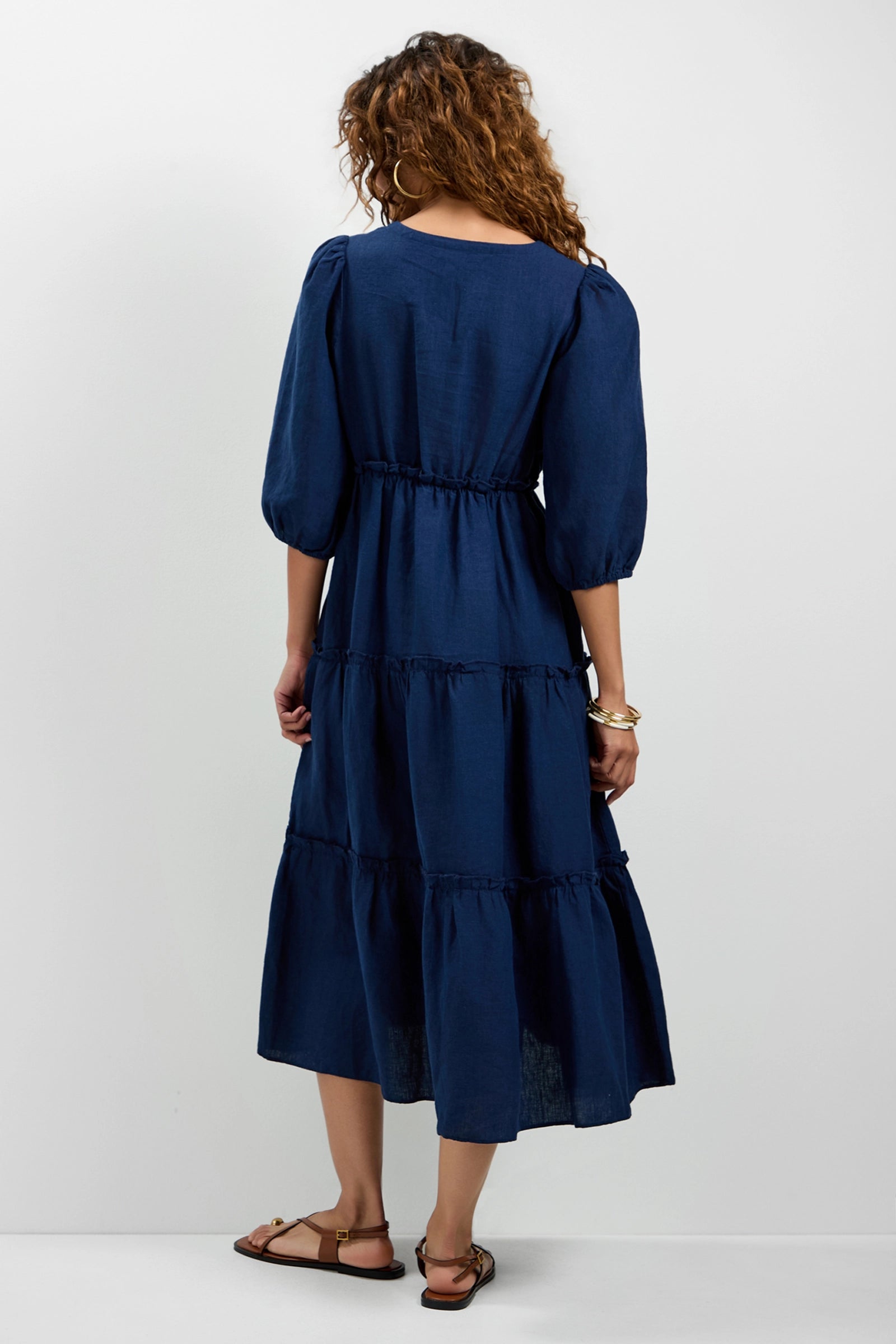 A woman with curly hair, wearing the Calm Tiered Ruffle Midi Dress in navy blue and brown sandals, stands facing away against a plain white background.