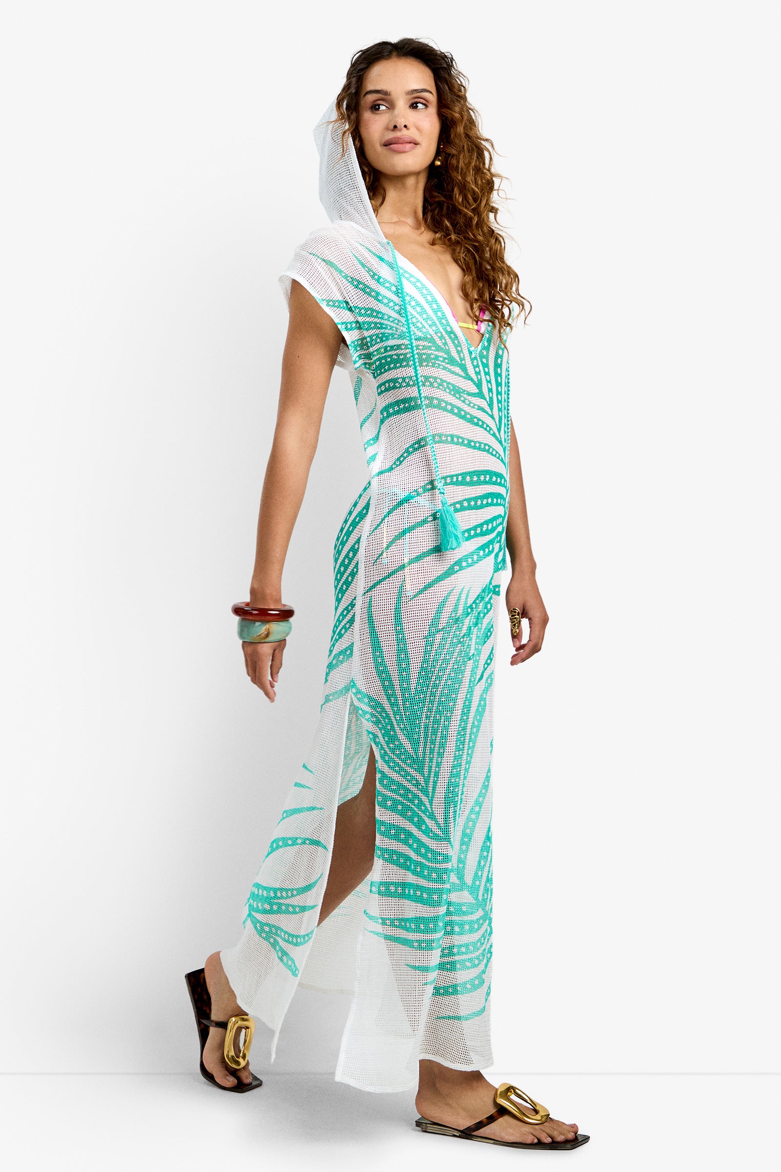 A woman wears the Cover-Up Maxi Dress - Sheer Tropics, a sheer white maxi with green palm leaf print, deep V-neck, hood, and side slit. She styles it with bangles, gold-accented flat sandals, long curly hair, and a pink bikini underneath.