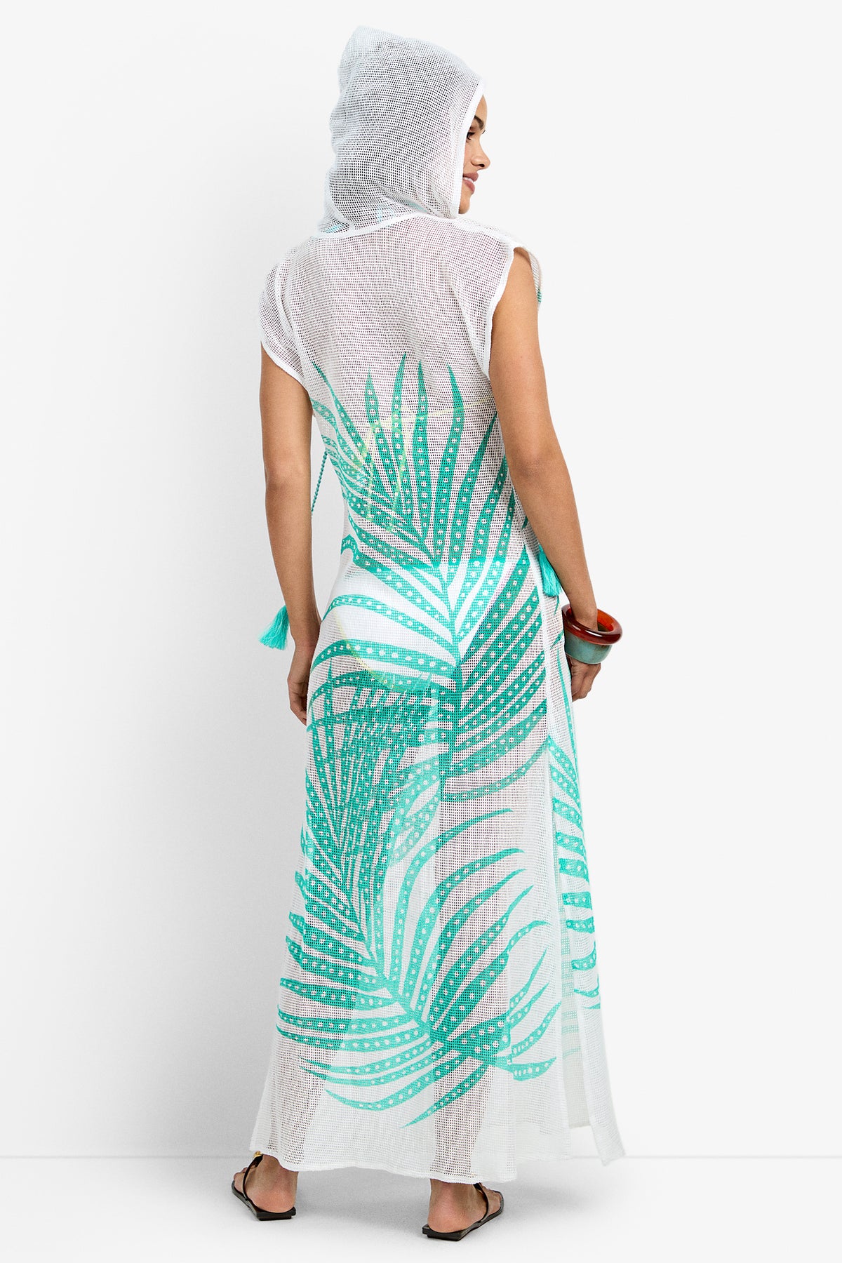 A barefoot person, shown from the back against a white background, wears the Cover-Up Maxi Dress - Sheer Tropics: a sleeveless, hooded, sheer white maxi dress with a bold green palm leaf print and holds a bag.