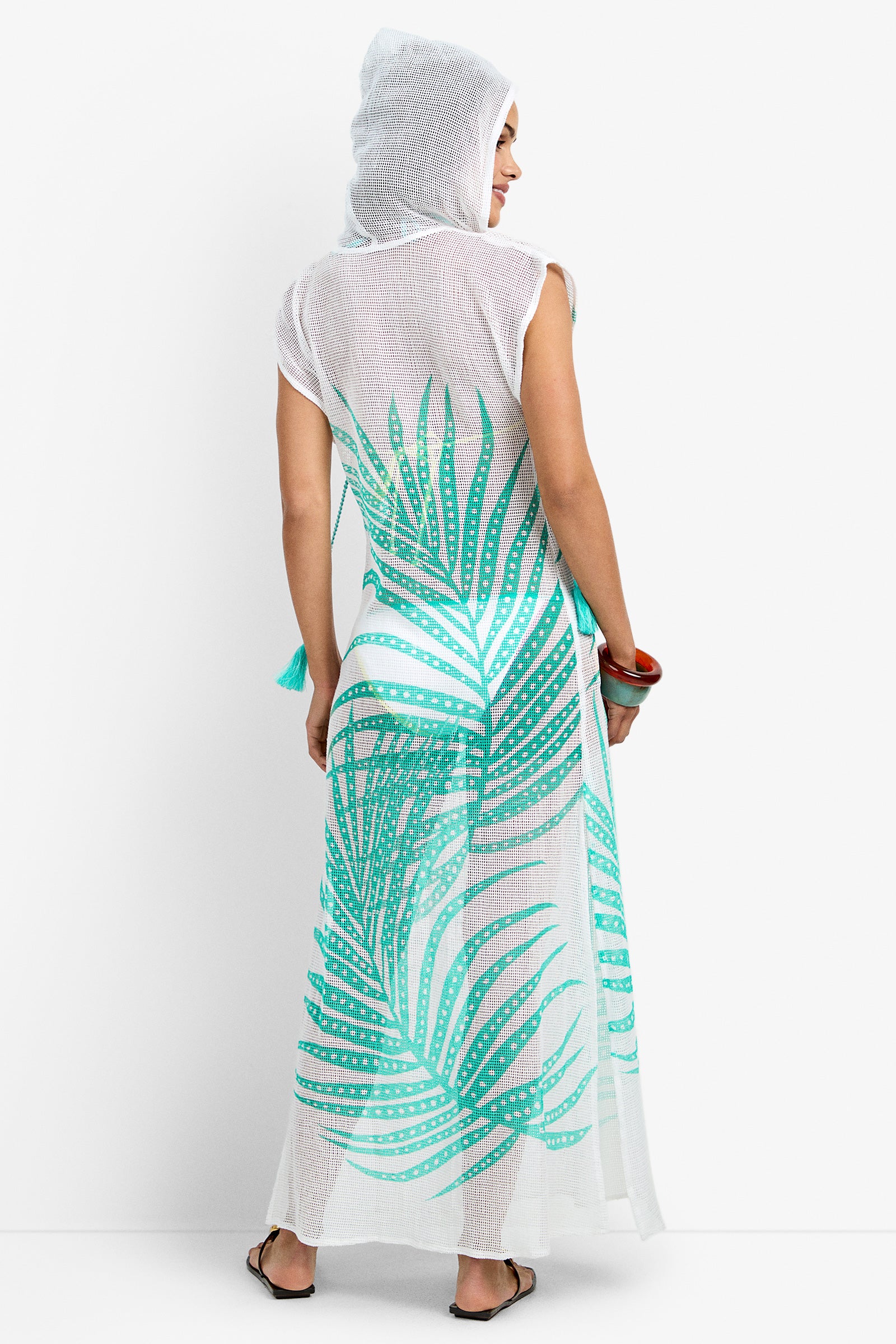 A barefoot person, shown from the back against a white background, wears the Cover-Up Maxi Dress - Sheer Tropics: a sleeveless, hooded, sheer white maxi dress with a bold green palm leaf print and holds a bag.