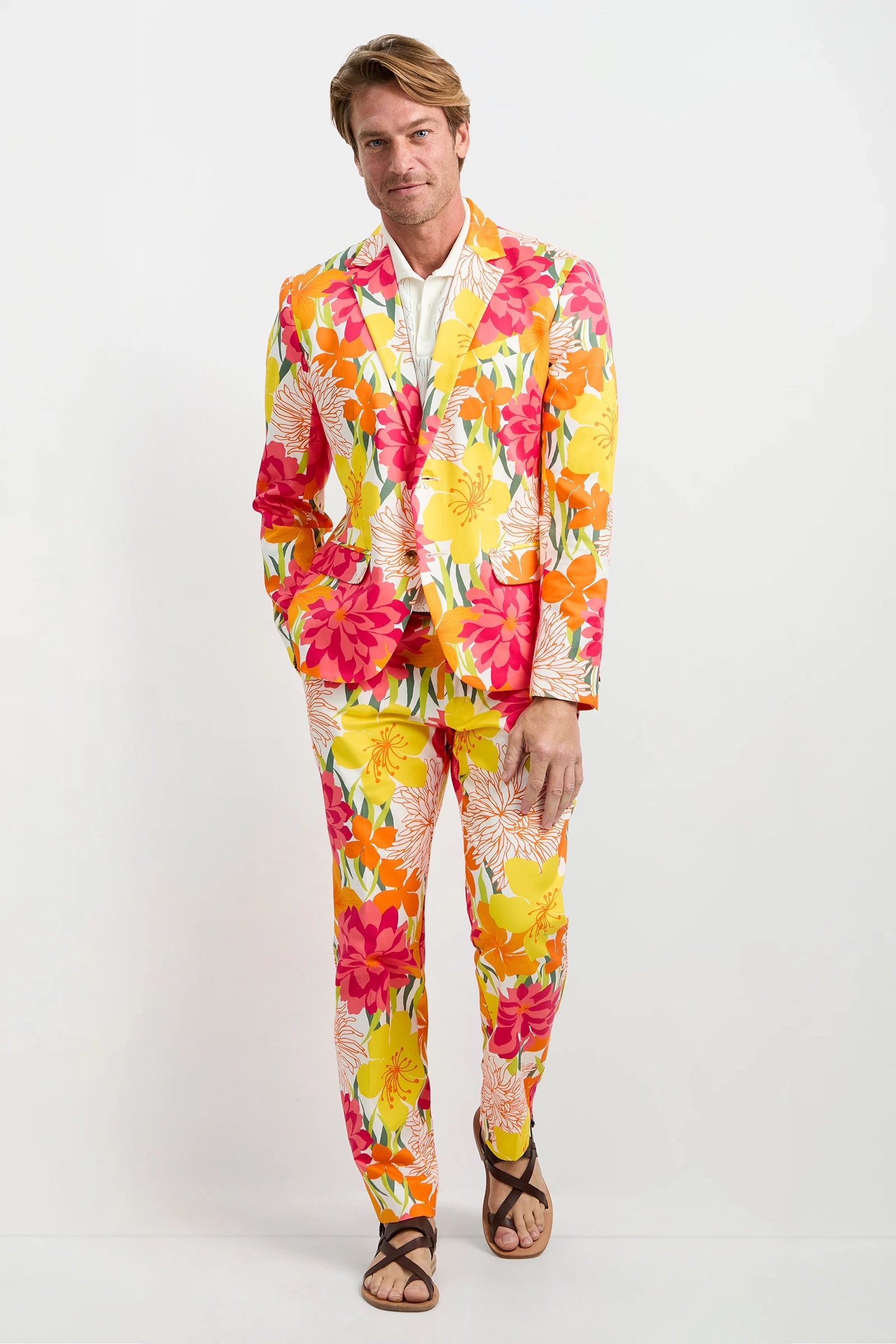 A man poses against a white background wearing the Thurston Blazer - Como Herringbone, paired with a white shirt and brown sandals.