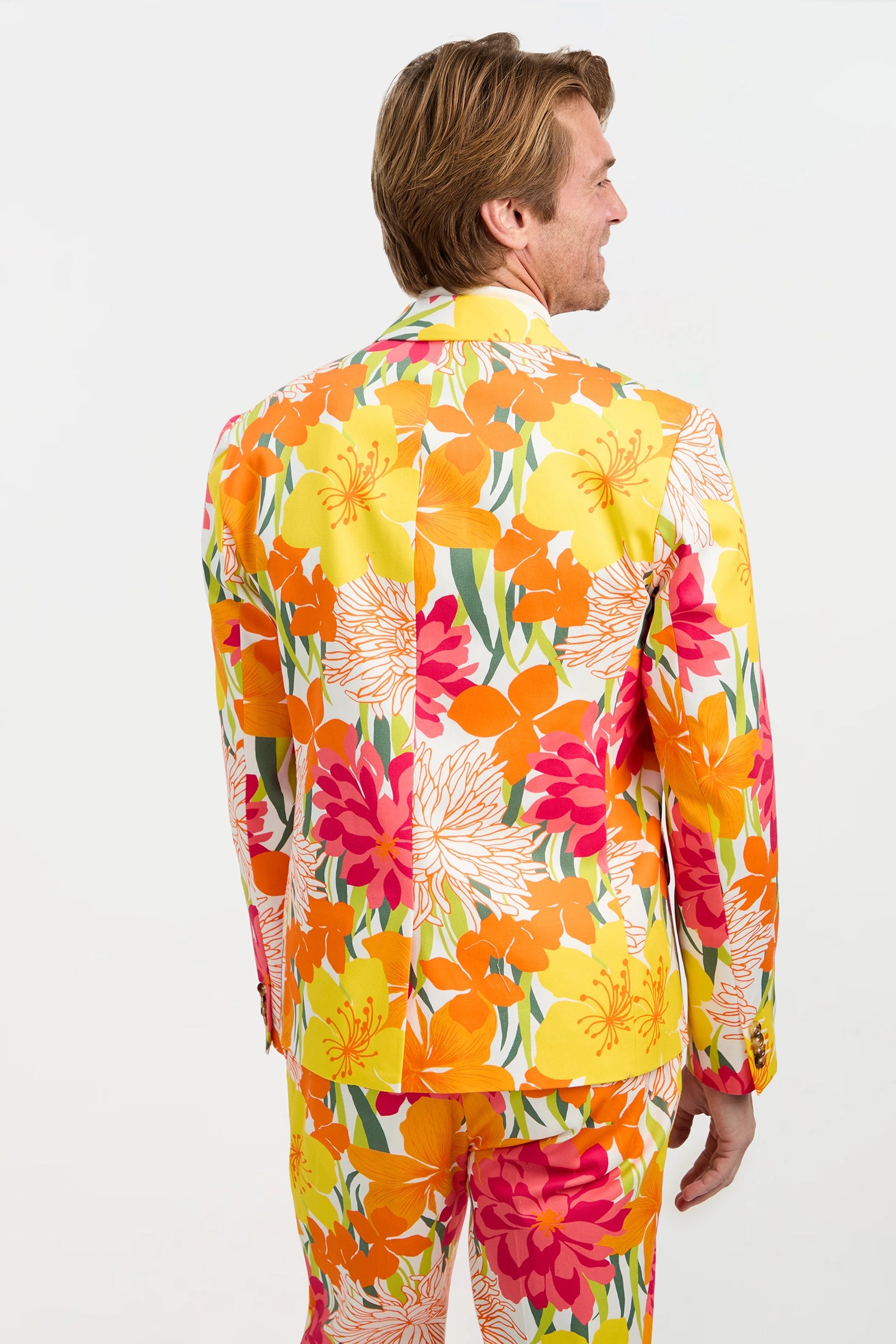 A man stands facing away, wearing the Thurston Blazer - Como Herringbone with a bold tropical floral pattern in yellow, orange, pink, and green against a plain white background.