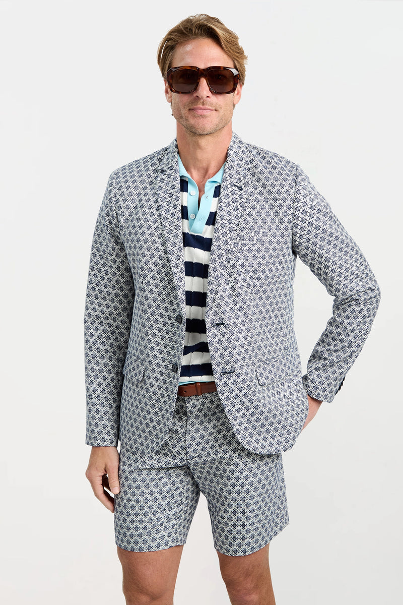 A man wearing the Thurston Blazer - Daisy Jacquard with matching shorts, a striped polo, sunglasses, and a brown belt stands against a plain white background.