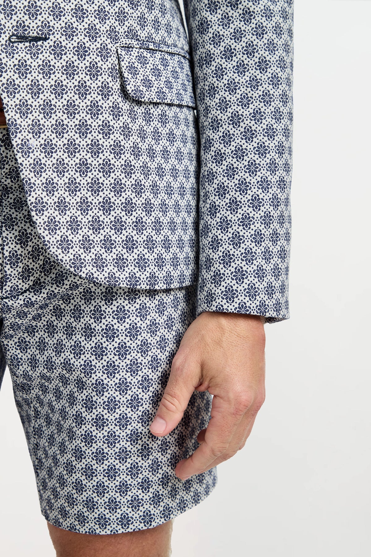 A person wearing the Thurston Blazer in Daisy Jacquard—a white and blue geometric patterned suit jacket and shorts—stands against a plain background with their right arm by their side.