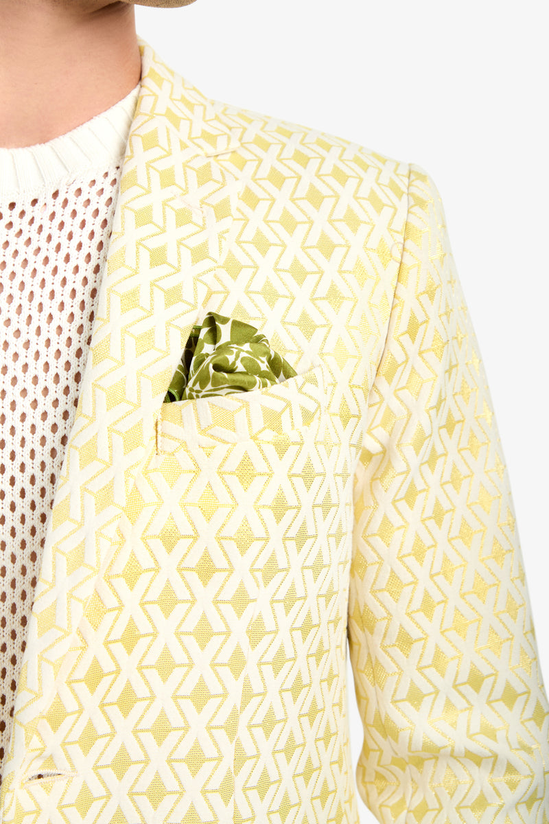 A close-up of a person wearing the Thurston Blazer - Crossroads Jacquard, featuring a yellow and white pattern, styled with a green pocket square and a white mesh-knit sweater. The background is plain and light-colored.
