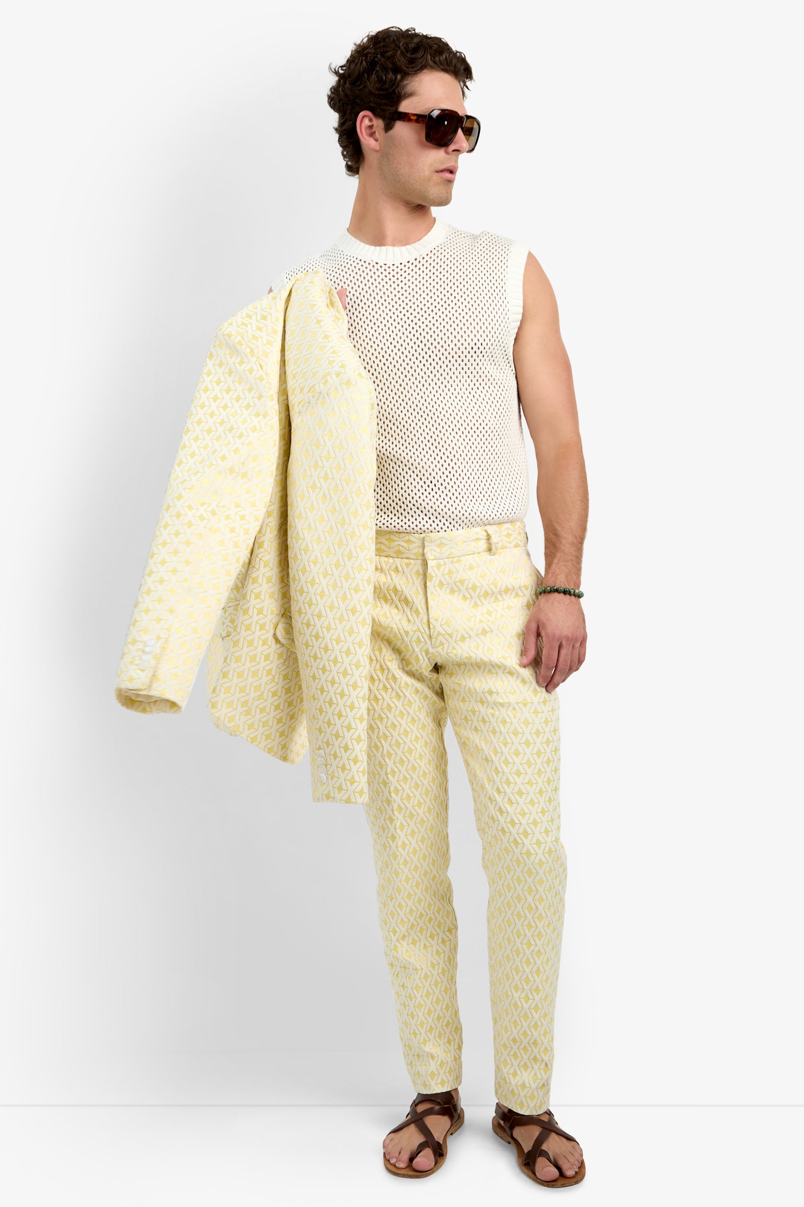 A man holds the Thurston Blazer - Crossroads Jacquard over his shoulder, wearing brown sunglasses, a sleeveless white mesh shirt, yellow patterned pants, and brown sandals while looking to the side.
