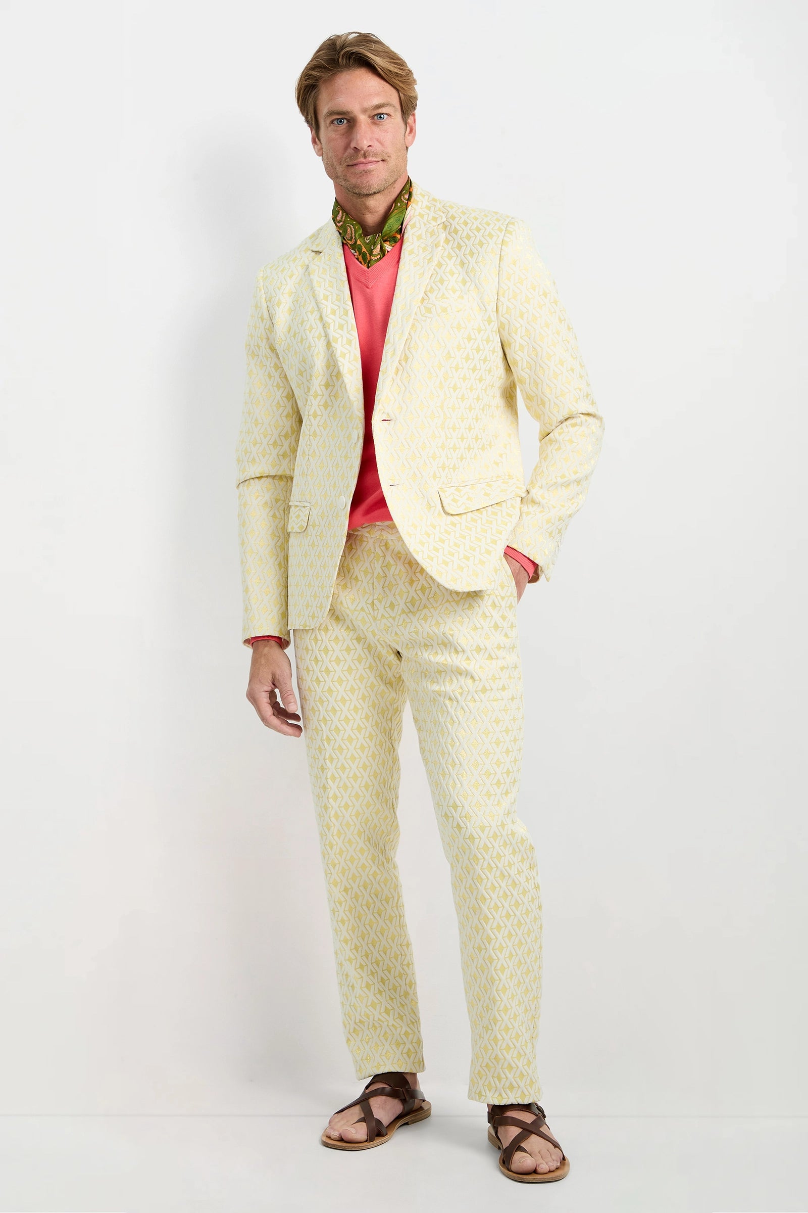 A man with light hair and a relaxed expression stands against a white background wearing the Clyde Slim Trouser - Crossroads Jacquard, paired with a pink sweater, green patterned scarf, and brown sandals.