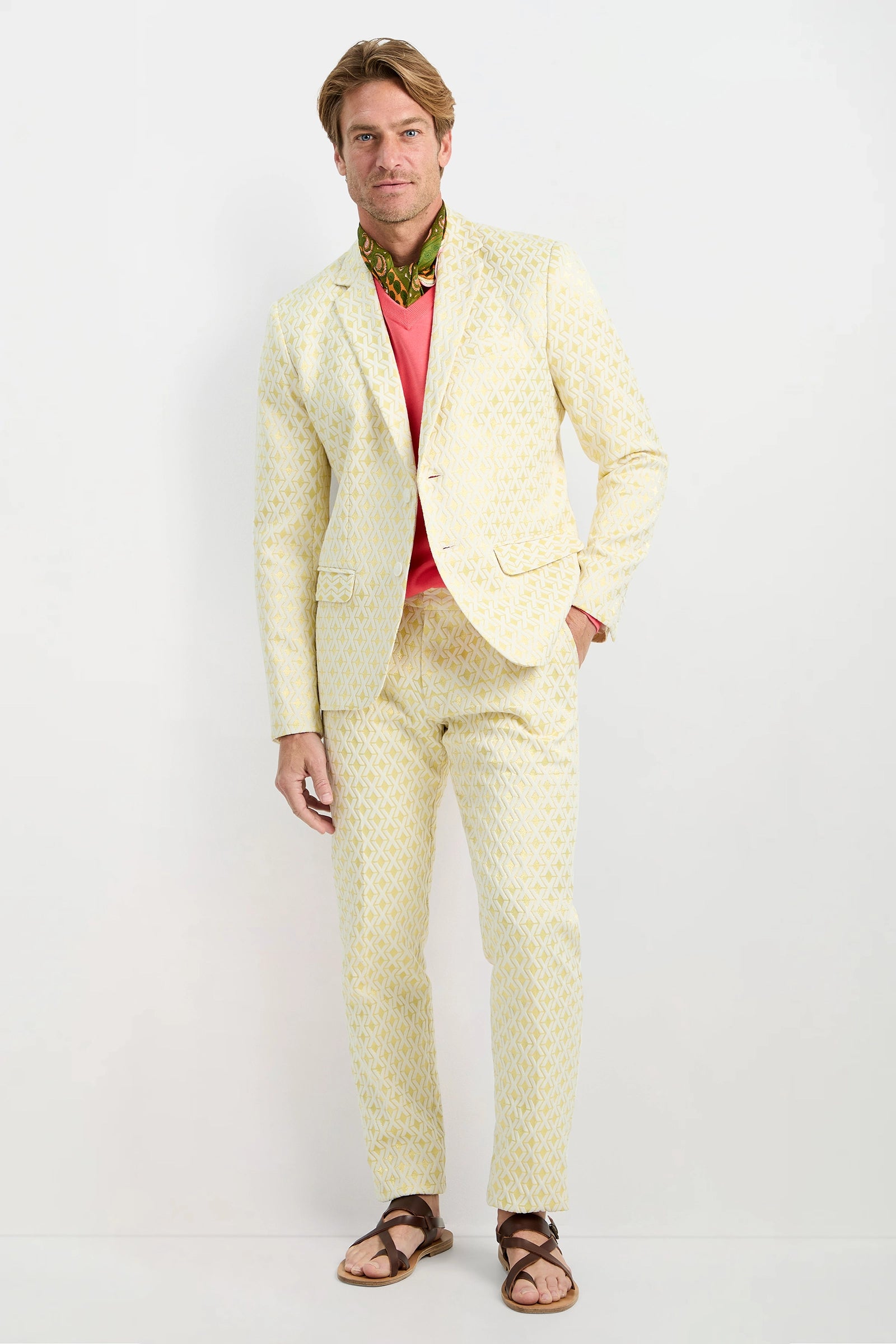 A man with light brown hair and a relaxed expression stands against a white background, wearing the Thurston Blazer in LEMON Crossroads Jacquard over a coral shirt, paired with a green patterned scarf and brown sandals.
