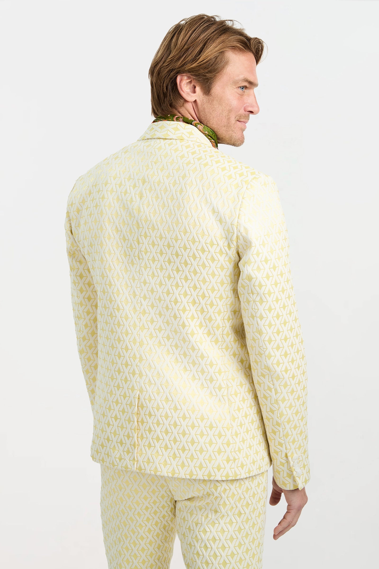 A man with light brown hair faces away, glancing over his shoulder. He wears the Thurston Blazer in Crossroads Jacquard, featuring a light yellow geometric pattern, paired with a green scarf against a plain white background.