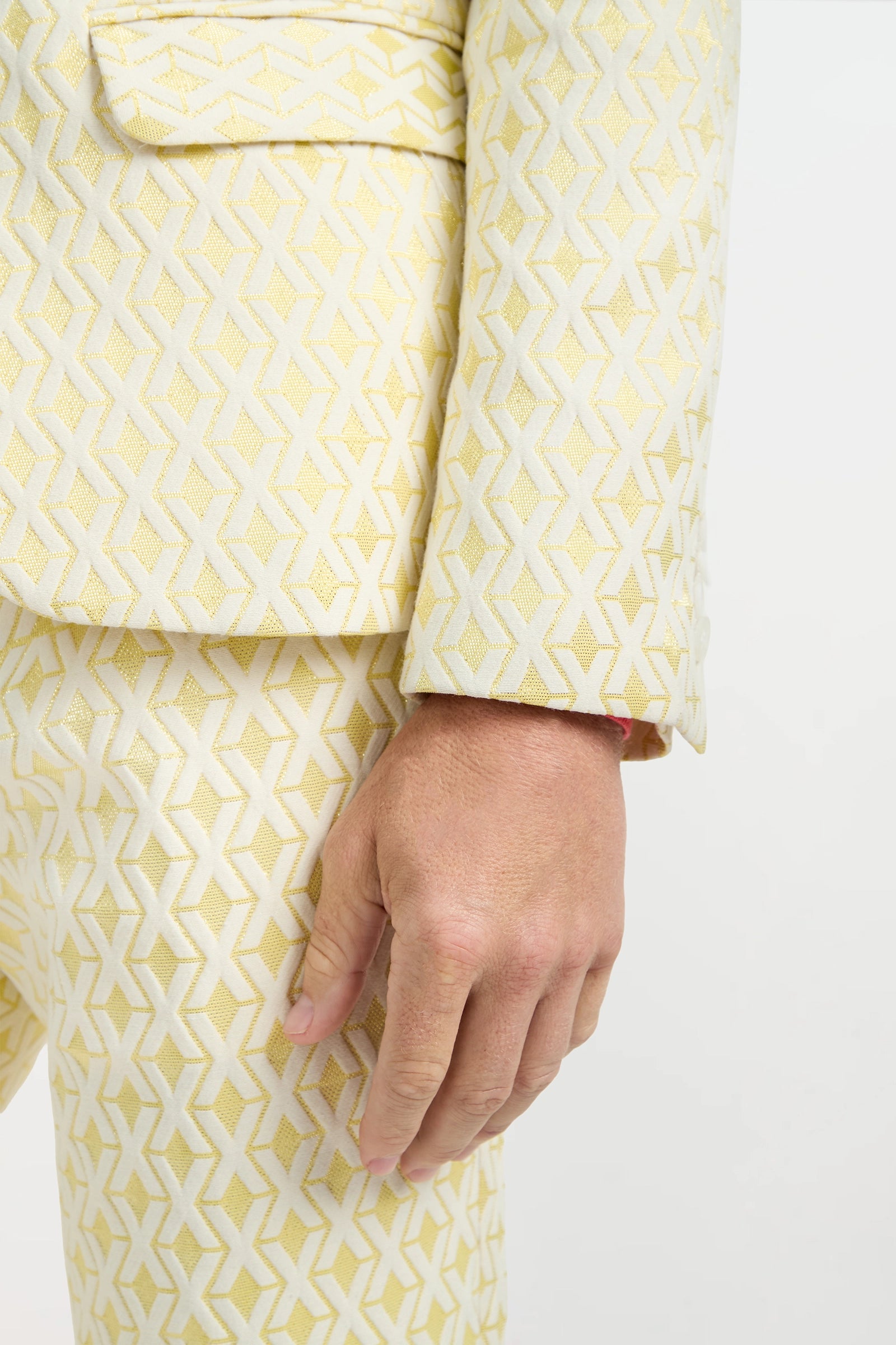 A close-up of a hand resting by the side, wearing the Thurston Blazer - Crossroads Jacquard in pale yellow with a white and gold geometric X pattern. The tailored blazer stands out against a plain white background.